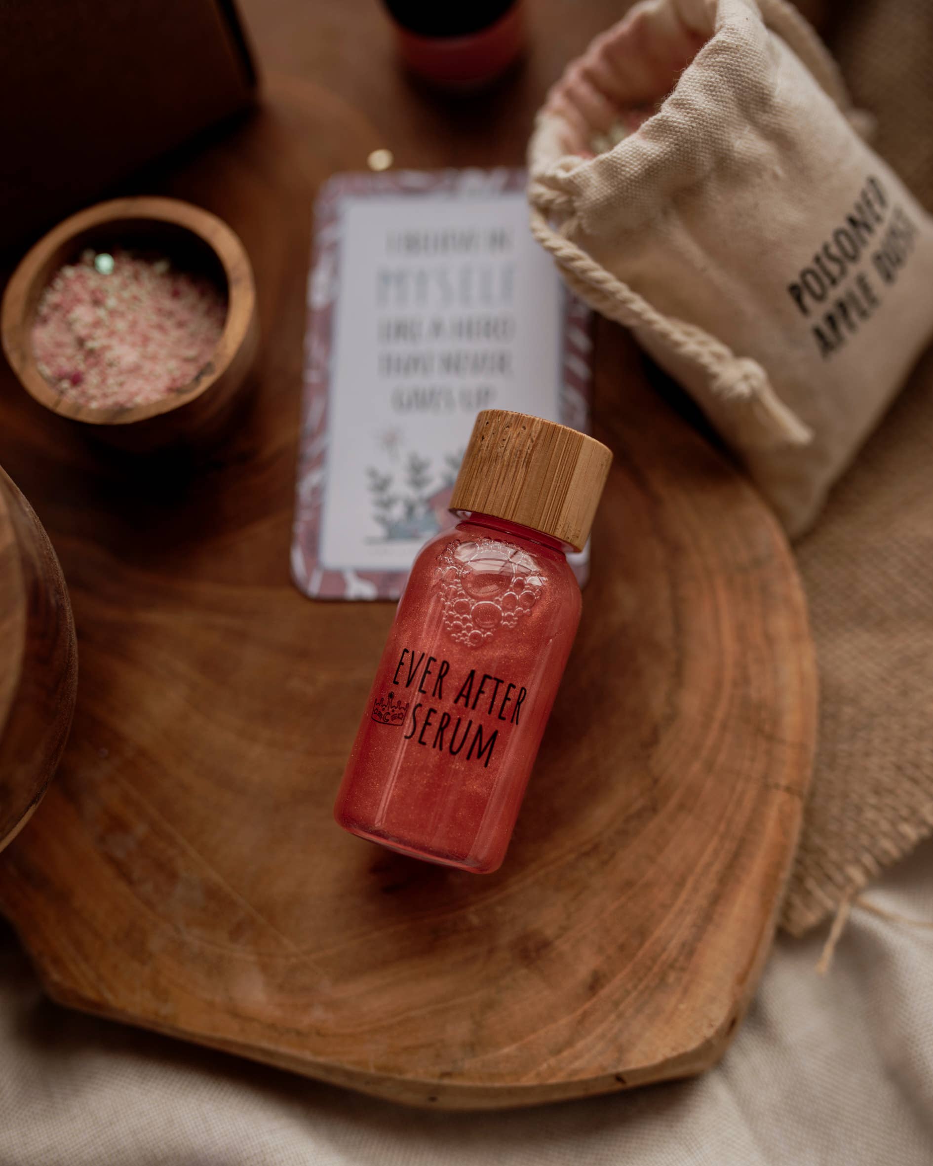A MINI Once Upon A Potion Kit by THE LITTLE POTION CO, featuring the "Ever After Serum" with a wooden cap, rests on wood. Nearby, a fabric pouch and packet of pink grains enhance its natural charm.