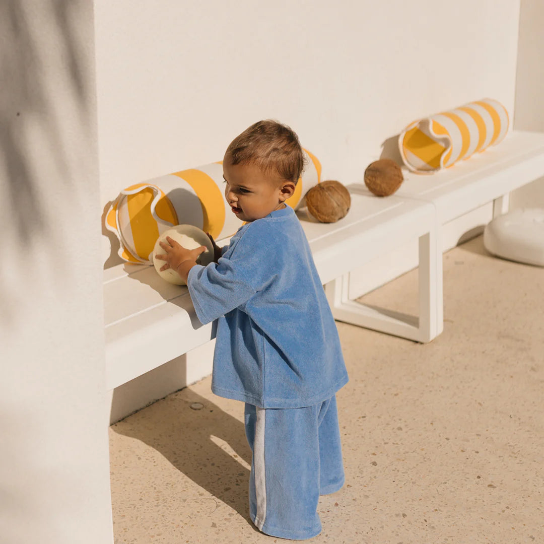 A child in GOLDEN CHILDREN Ciao Tutti relaxed pants (Terry Towel Pool Blue) stands by a white bench holding a coconut. The sunny setting is adorned with two striped yellow and white cushions and coconuts, featuring a light-colored floor and wall, creating an inviting outdoor scene.