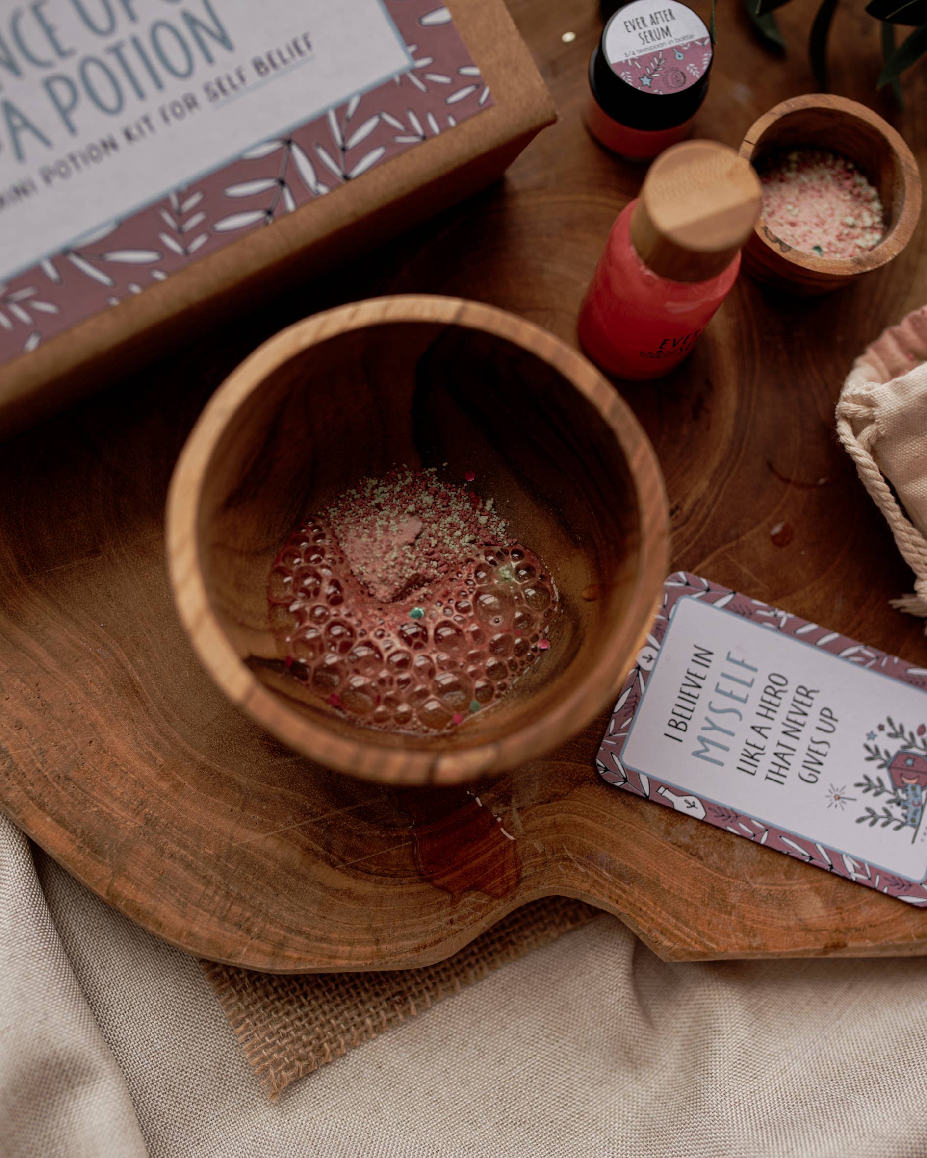 A wooden cup with colorful powder and bubbles sits on a wooden surface, creating an enchanting vibe. Nearby are small bottles and a card saying "Believe in Myself." The Little Potion Co's MINI Once Upon A Potion Kit (a potion for self belief) is partially visible in warm, earthy tones.