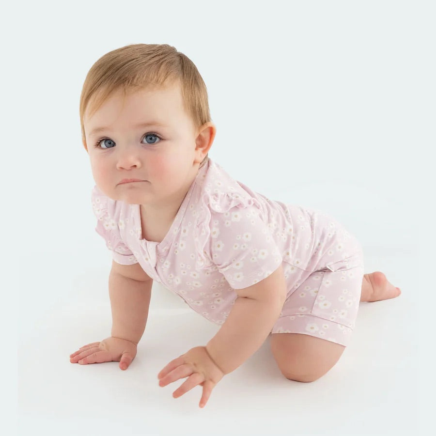 A baby with light brown hair crawls on a pale surface, wearing KYND BABY's Day or Night Short Sleeve Frill Onesie in Orchid Bloom, featuring small white daisy patterns. The sustainable fabric enhances comfort as they gaze upward.