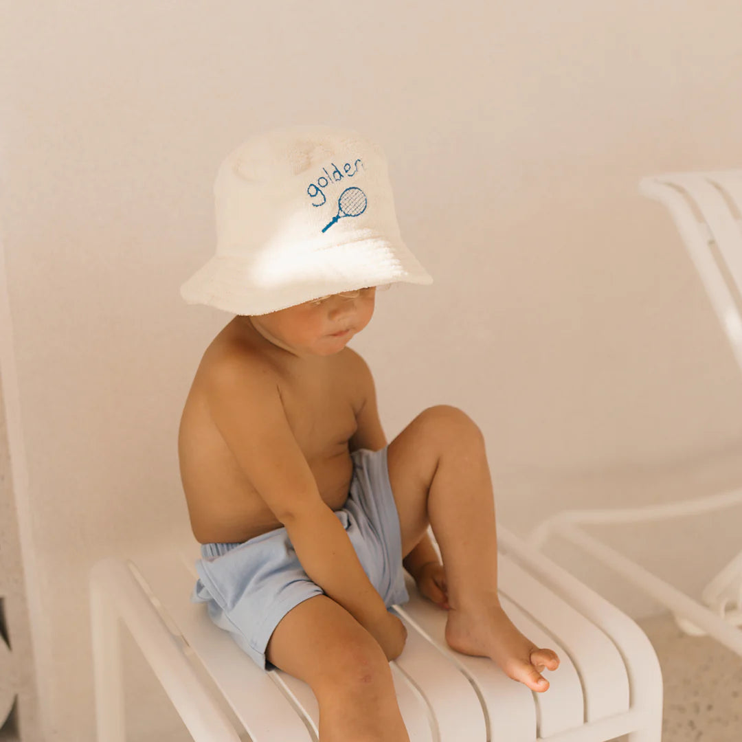 A small child sits on a white chair, wearing a white hat with a tennis racket graphic and "golias," and Retro Boy Shorts Palm Blue by GOLDEN CHILDREN. Against the plain white background, they embody a relaxed fit with their neutral expression.