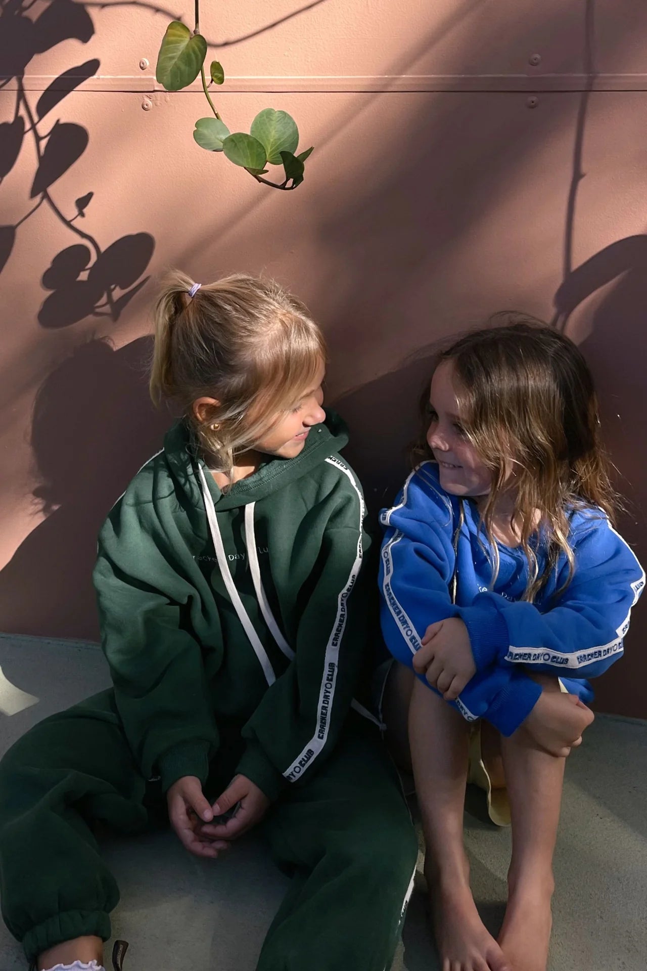 Two young children sit on the ground, smiling at each other. One wears a green tracksuit, while the other sports an oversized CRACKER DAY CLUB Kids Cloud Racer Hoodie Forest. Sunlight casts leafy shadows on the pink wall behind them.