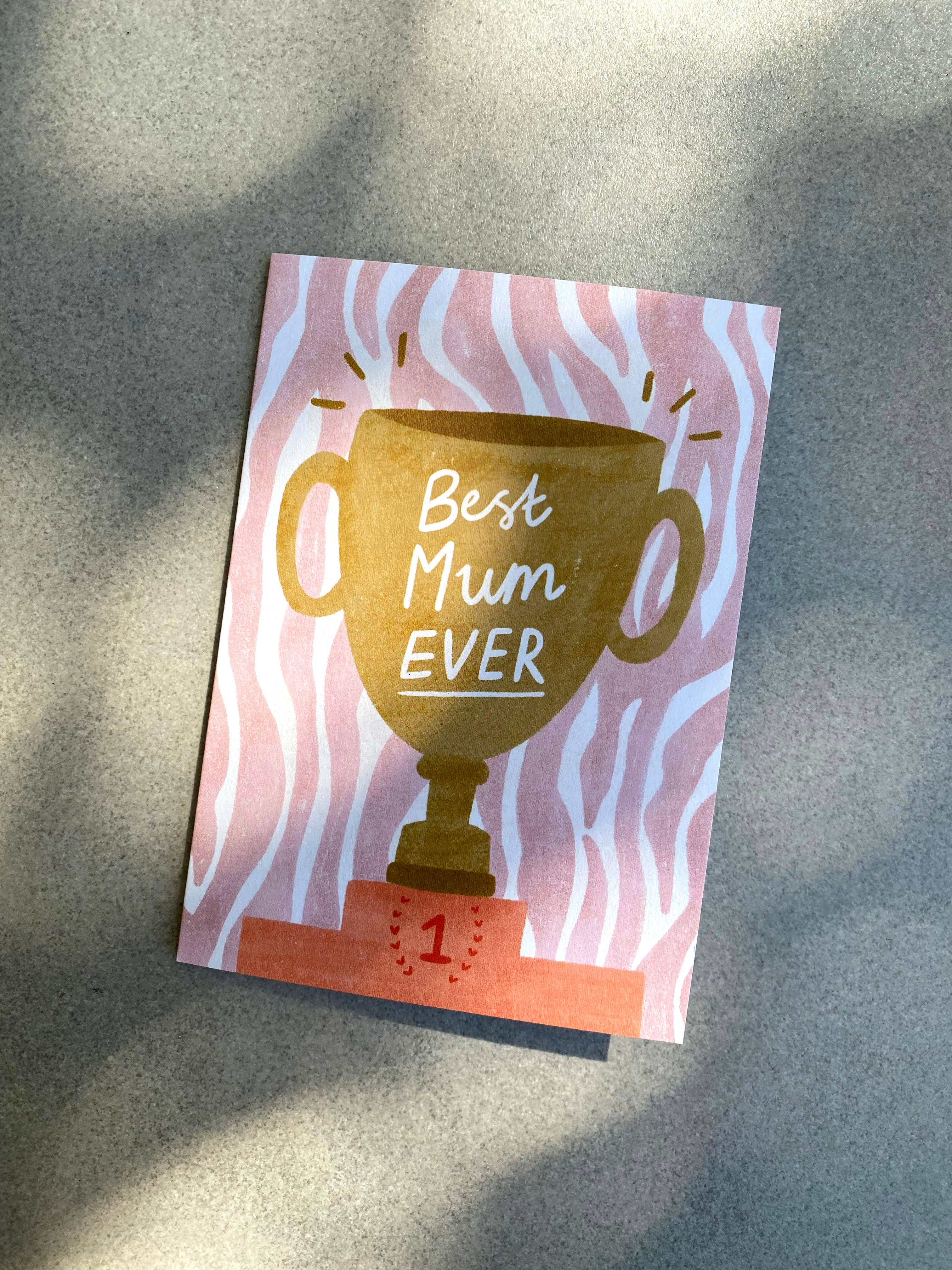 Celebrate your amazing mum with the "Best Mum Ever Trophy Greeting Card" by Lauren Sissons Studio, featuring a gold trophy with “Best Mum Ever” and a “1” on the base, set on a pink and white wavy background. Made from recycled materials.