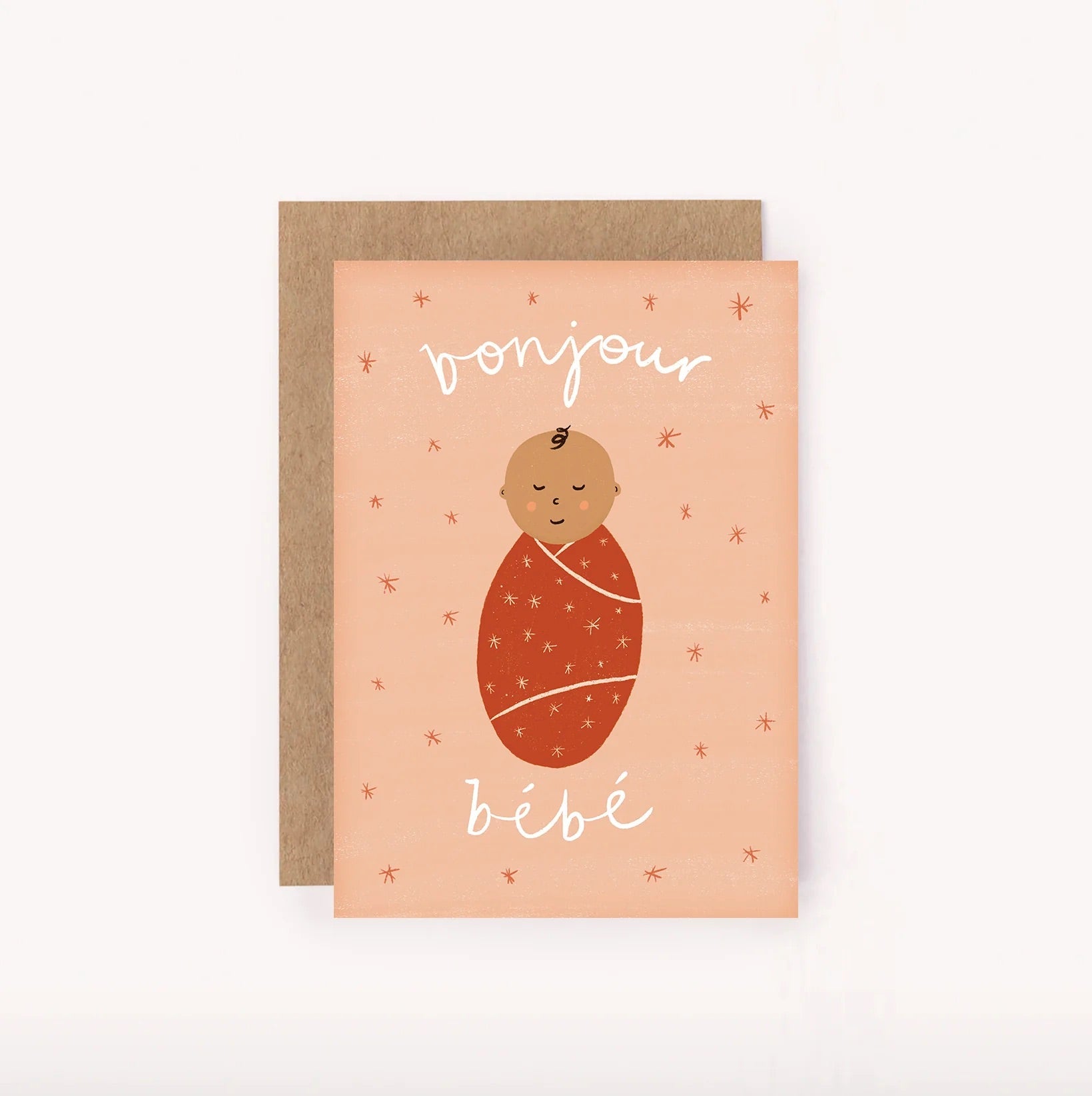 The "Bonjour Bebe Mini Card Pink" from LAUREN SISSONS STUDIO showcases a charming illustration of a smiling baby wrapped in a red-orange blanket against a peach background adorned with star-like accents. This recycled paper mini greeting card, perfect for baby showers, is accompanied by a brown envelope tucked neatly behind it.