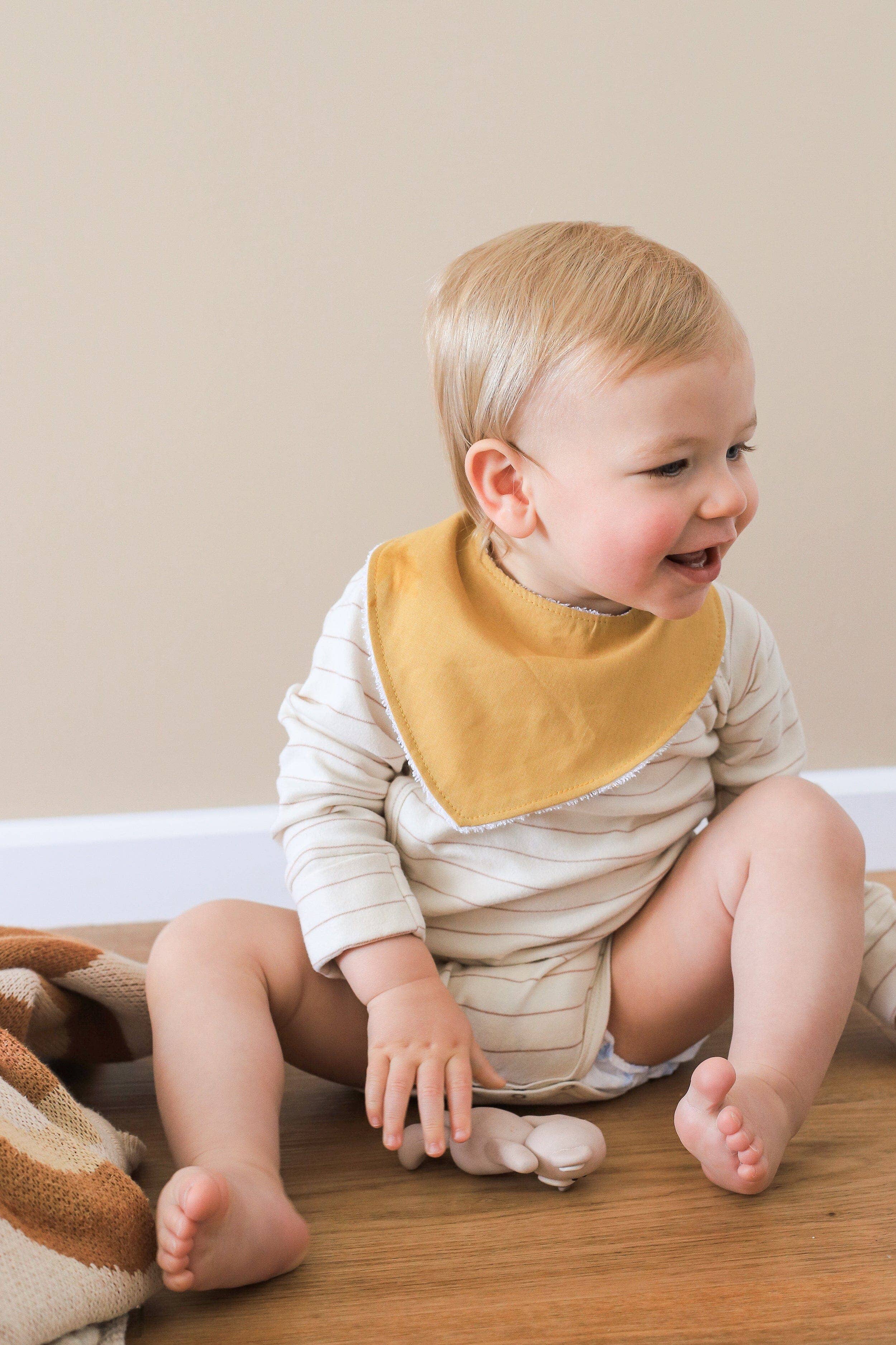 A happy blonde baby sits on the floor wearing a striped onesie and a Kiin Baby Dribble Bib made of absorbent bamboo/cotton. Holding a small toy, the baby is near a knitted blanket against a plain, light-colored wall.