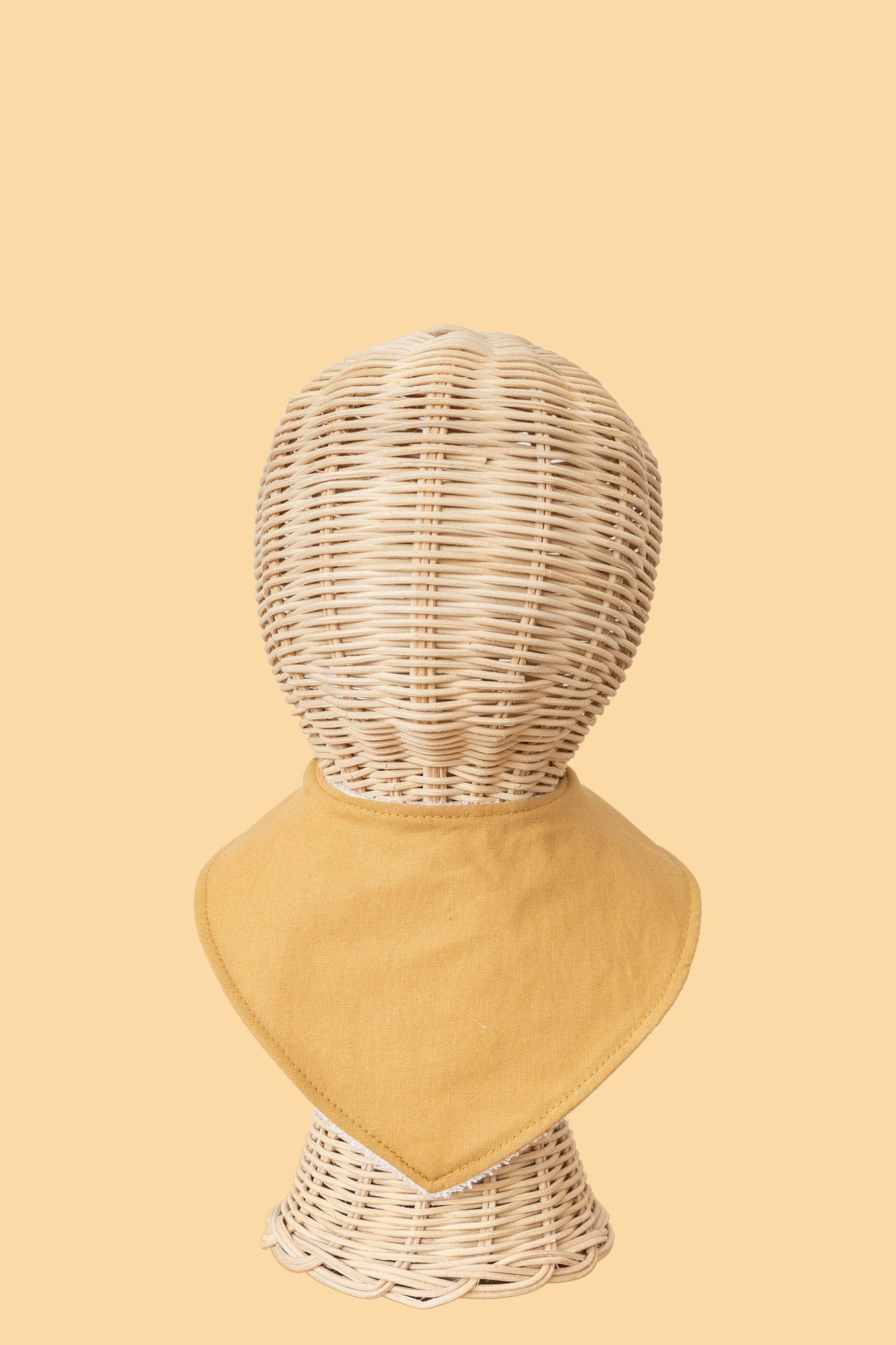 A wicker mannequin head is elegantly adorned with Kiin Baby's Dribble Bib, a mustard yellow bandana crafted from a soft bamboo/cotton blend against a light yellow backdrop.