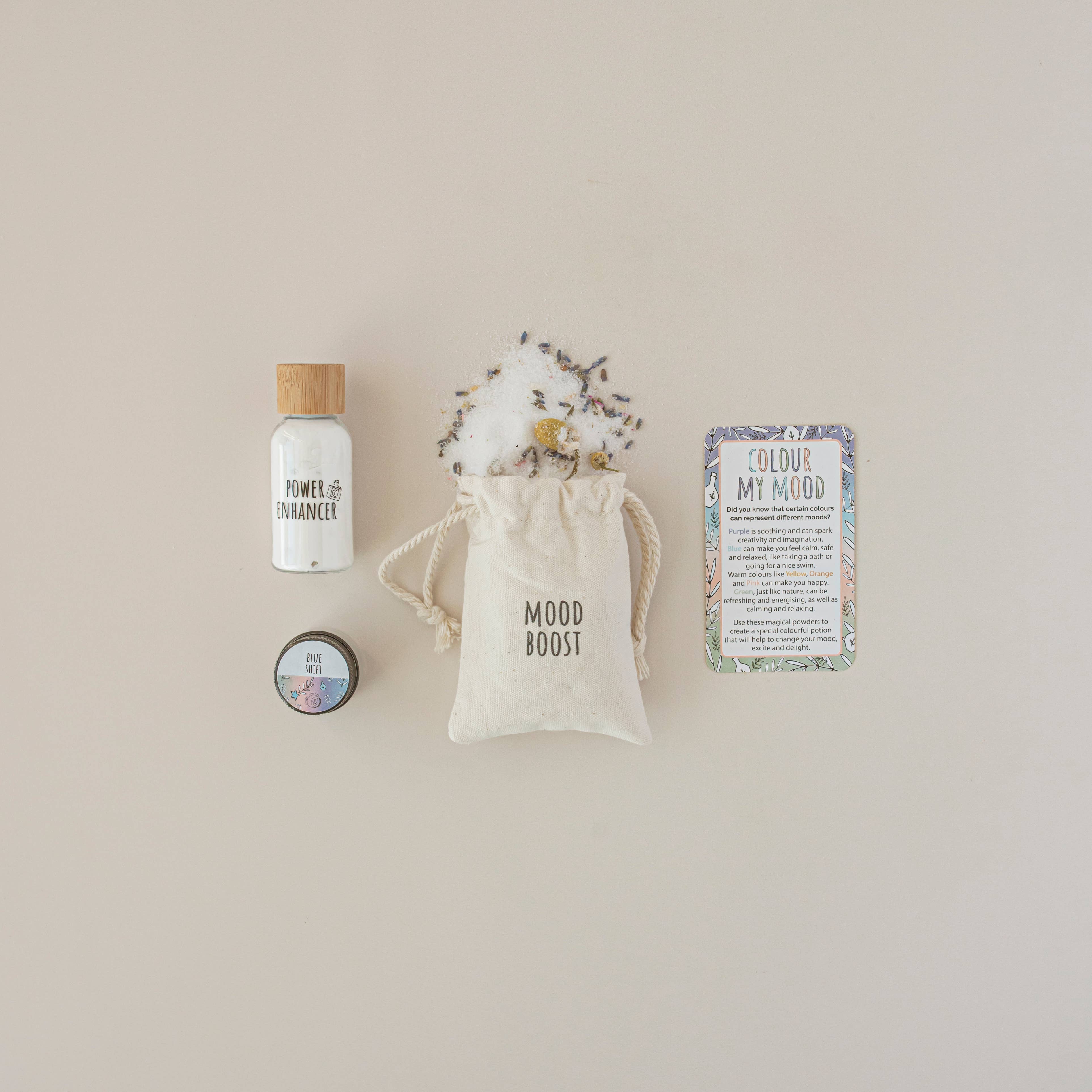 A flat lay of The Little Potion Co's MINI Colour Mood Potion Kit (Colour Changing Kit) for sensory play showcases a "Power Enhanced" bottle, a "Mood Boost" herb pouch, a "Blue Tansy Clay" jar, and a "Colour My Mood" card on beige, designed to inspire mood change.