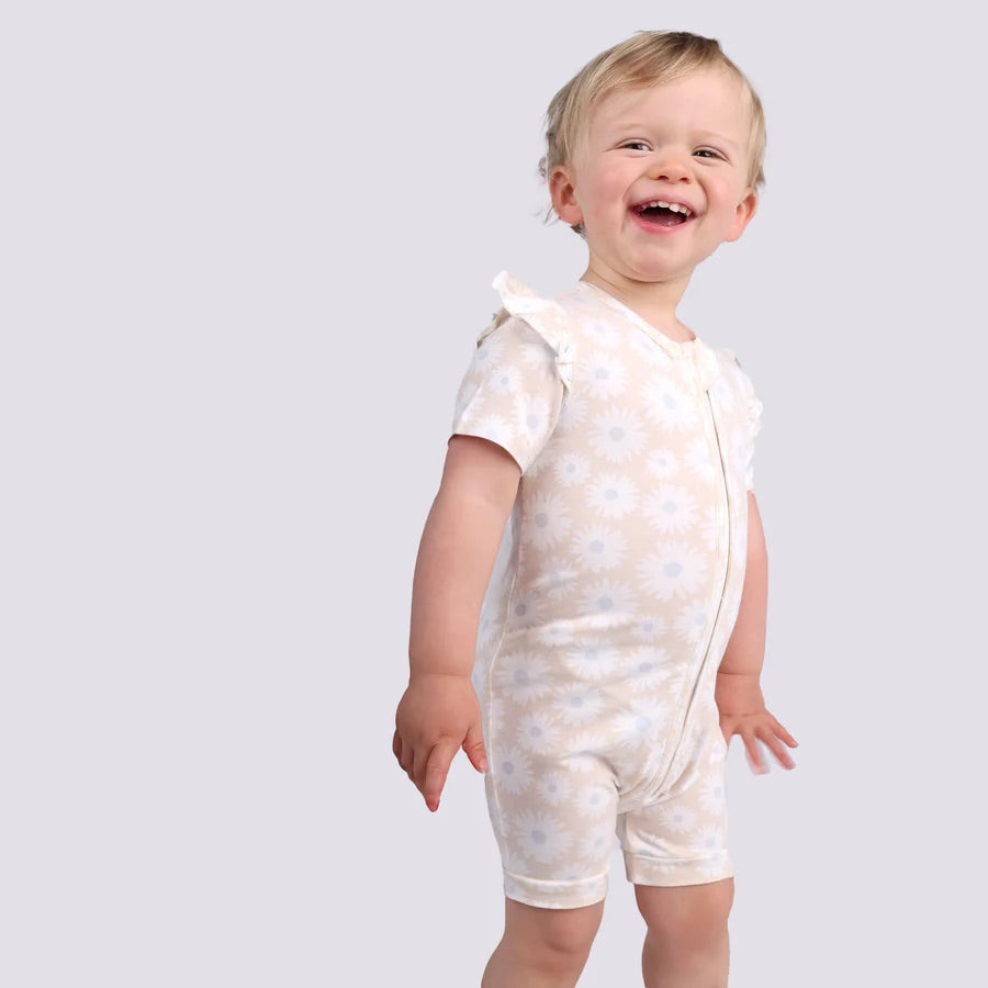 A content toddler stands energetically against a plain background in KYND BABY's Day or Night Short Sleeve Frill Onesie Petal Fields, made from EcoCosy Viscose. The light floral romper with ruffles offers practical comfort for summer.