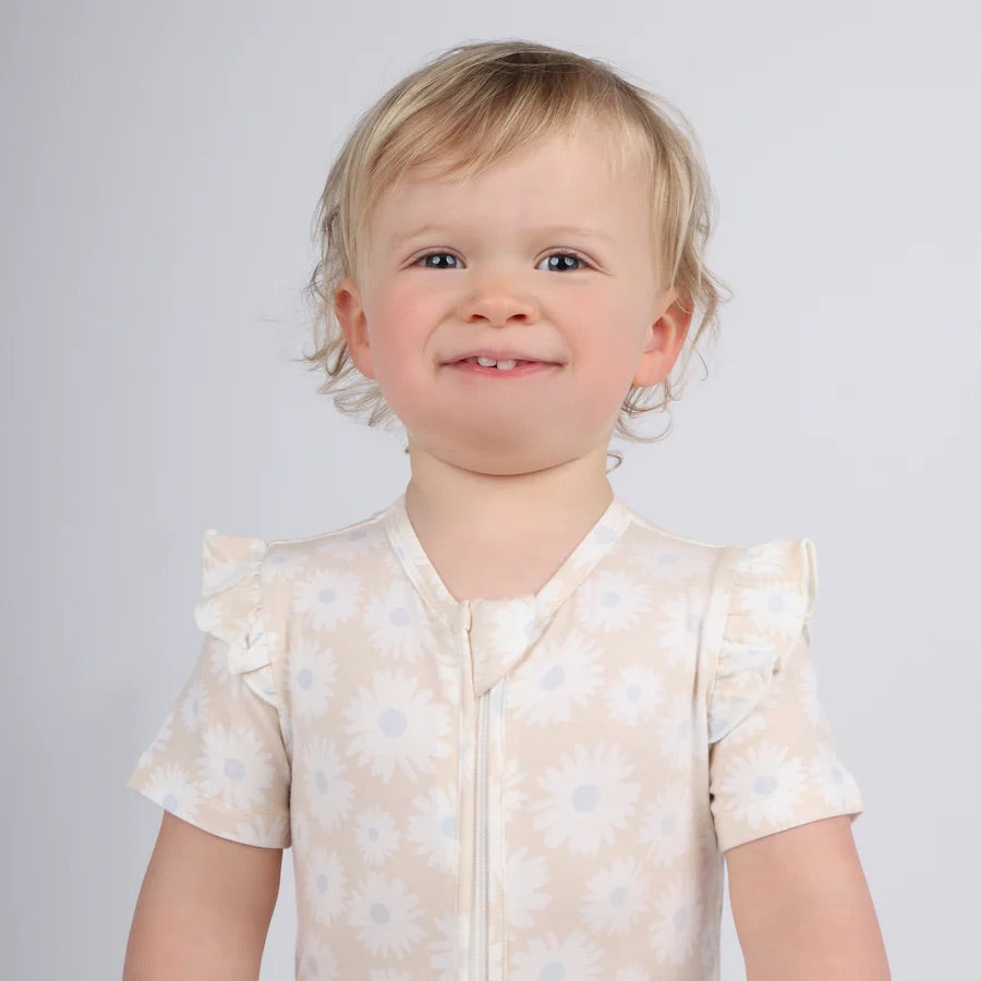 A toddler with short, light hair beams happily in a KYND BABY Day or Night Short Sleeve Frill Onesie Petal Fields, featuring daisies and ruffled sleeves, against a plain, light gray background—showcasing summer practicality in style and comfort.