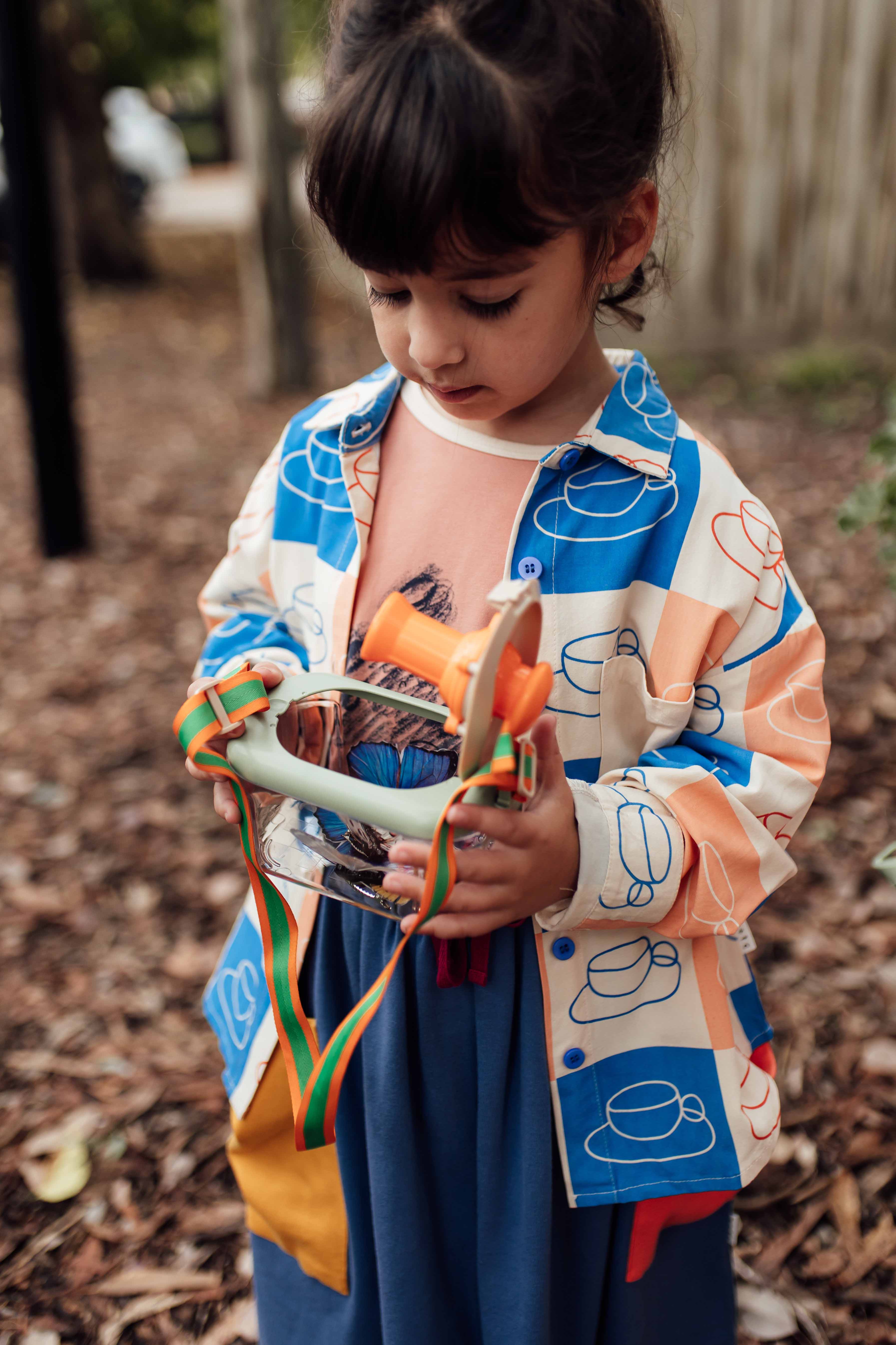A young girl with dark hair is outdoors, dressed in a colorful Cups Button Down Shirt by OLIVE + THE CAPTAIN and a blue skirt. She is attentively looking down at an object she's holding, which appears to be a small hand drum with ribbons attached. The background features mulch and some trees.