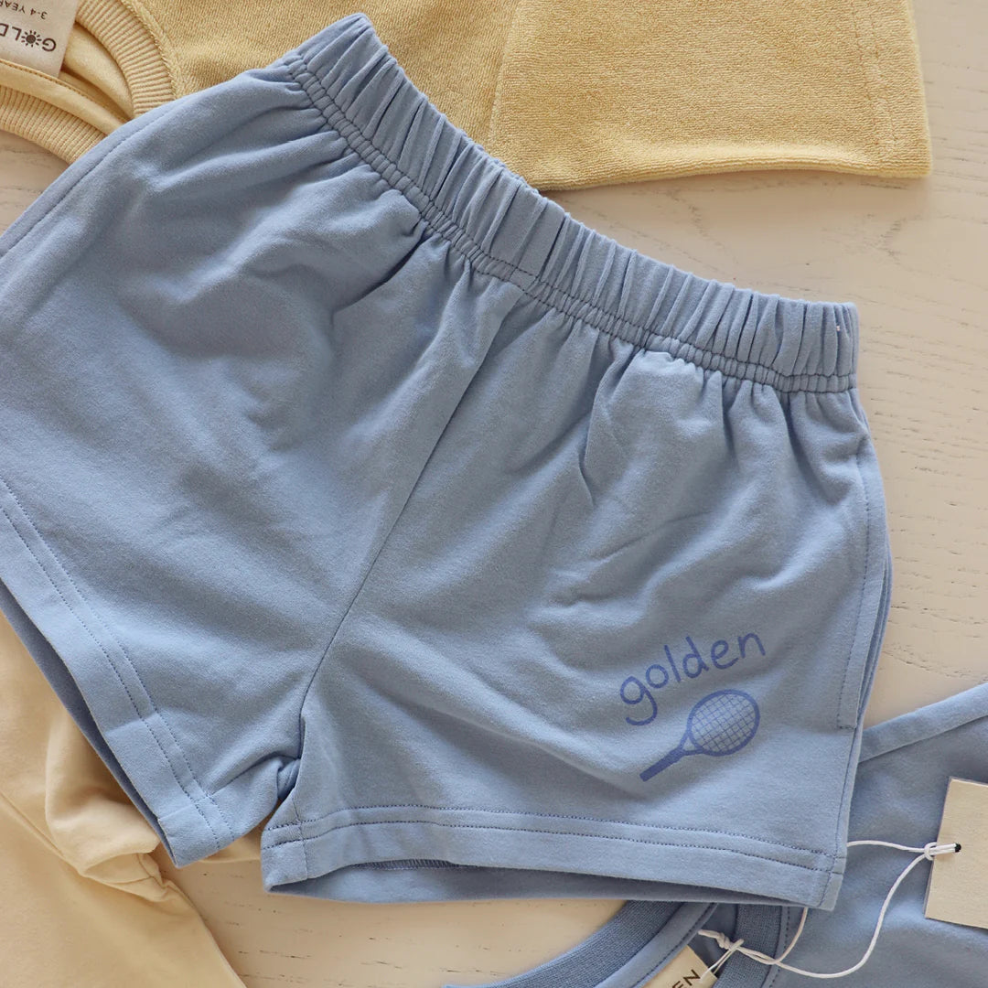 Palm Blue Retro Boy Shorts by GOLDEN CHILDREN, made from cotton jersey, feature a relaxed fit with an elastic waistband and display a tennis racket graphic beside "golden." They lay on a light surface next to beige clothing.