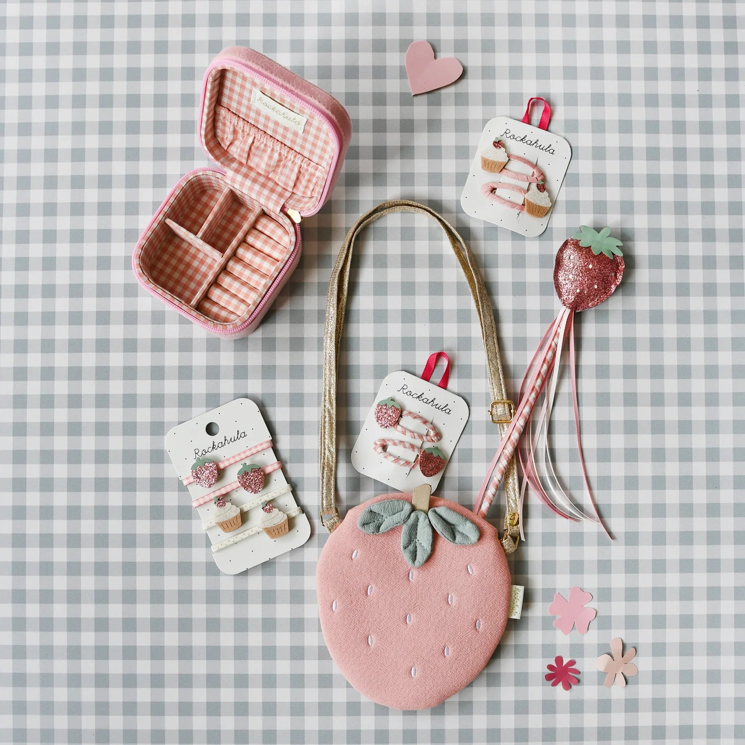 Flat lay of adorable strawberry-themed accessories on a gray plaid background, featuring the ROCKAHULA Sweet Strawberry Square Jewellery Box, a pink strawberry purse, hair clips, a glittery strawberry wand, and pink flower cutouts.
