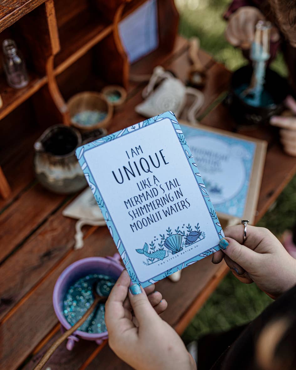 A person is holding a card adorned with decorative blue borders that reads, "I am unique like a mermaid's tail shimmering in moonlit waters." In the background, a wooden shelf displays small bottles from THE LITTLE POTION CO's Moonlight Waters MINDFUL Potion Kit, while a hand stirs a blue substance in a bowl—ideal for sensory play and mindfulness.