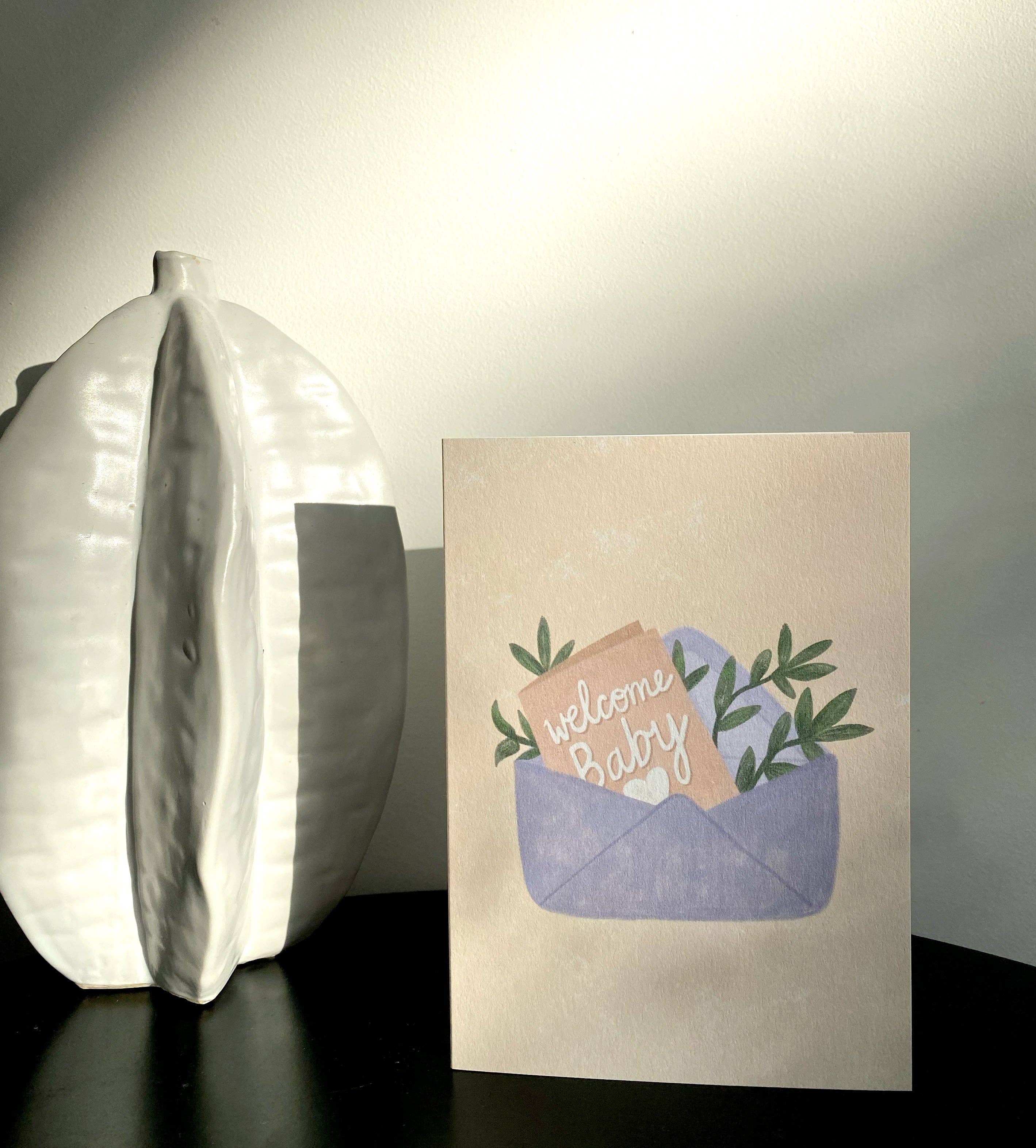 A "Welcome Baby" greeting card by Lauren Sissons Studio, gender-neutral and crafted from recycled paper, features a baby item illustration in a blue basket on a dark surface. Alongside it, a white ceramic vase with textured design casts gentle shadows across the scene.