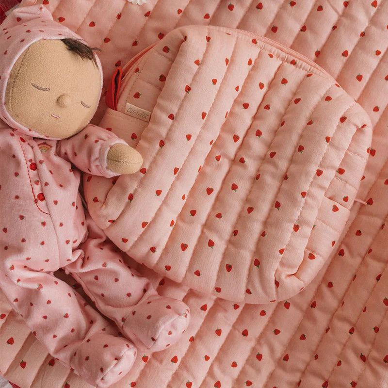 A soft doll in a pink onesie with red hearts rests beside the OLLI ELLA Quilted Toiletries Bag Strawberry, both set on a matching pink quilted blanket with red hearts.
