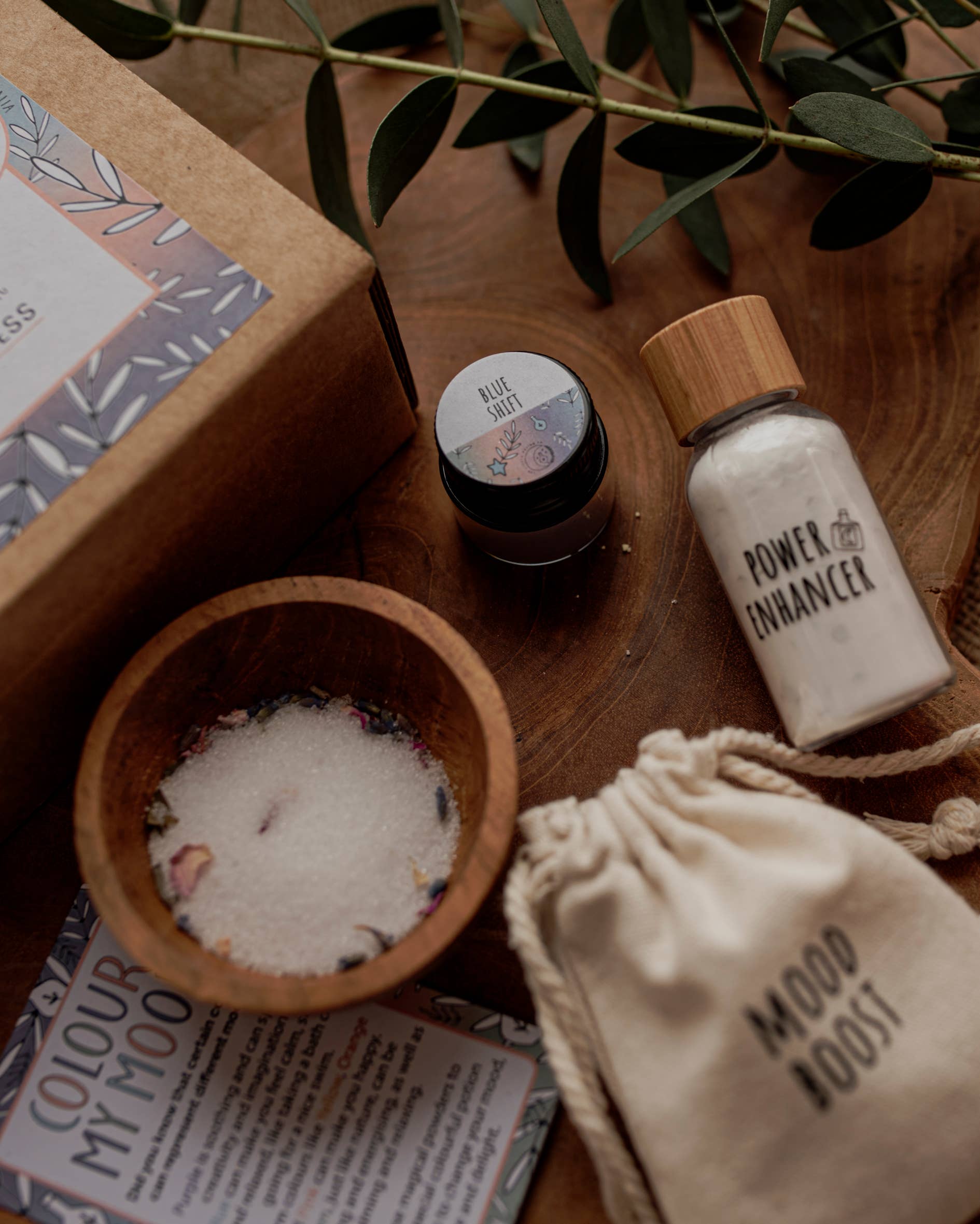 A flat lay displays The Little Potion Co's MINI Colour Mood Potion Kit (Colour Changing Kit) on a wooden surface, featuring "Blue Bath" jar, "Power Enhancer" bottle, "Mood Boost" sensory play pouch, accompanied by bath salts and a "Colour My Mood" card suggesting mood changes.