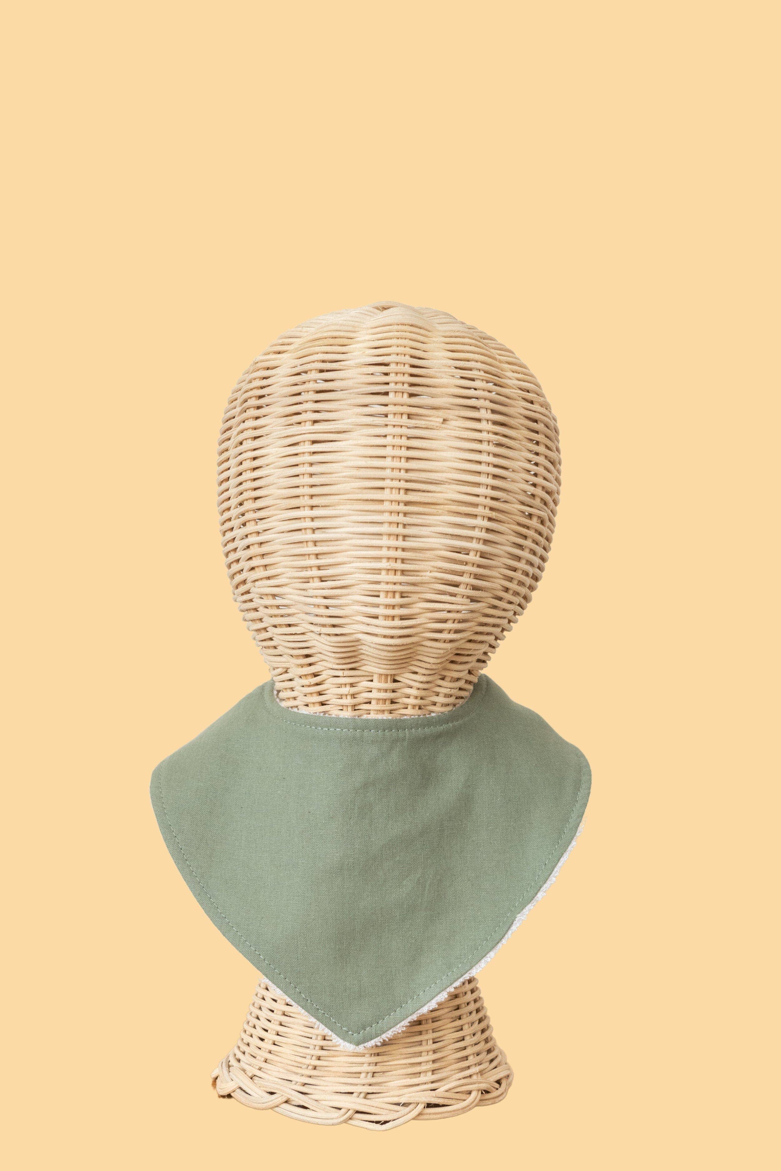 A woven rattan mannequin head displays a Kiin Baby Dribble Bib, featuring a sage green bamboo/cotton fabric bandana, set against a soft beige background for stylishly absorbent baby functionality.