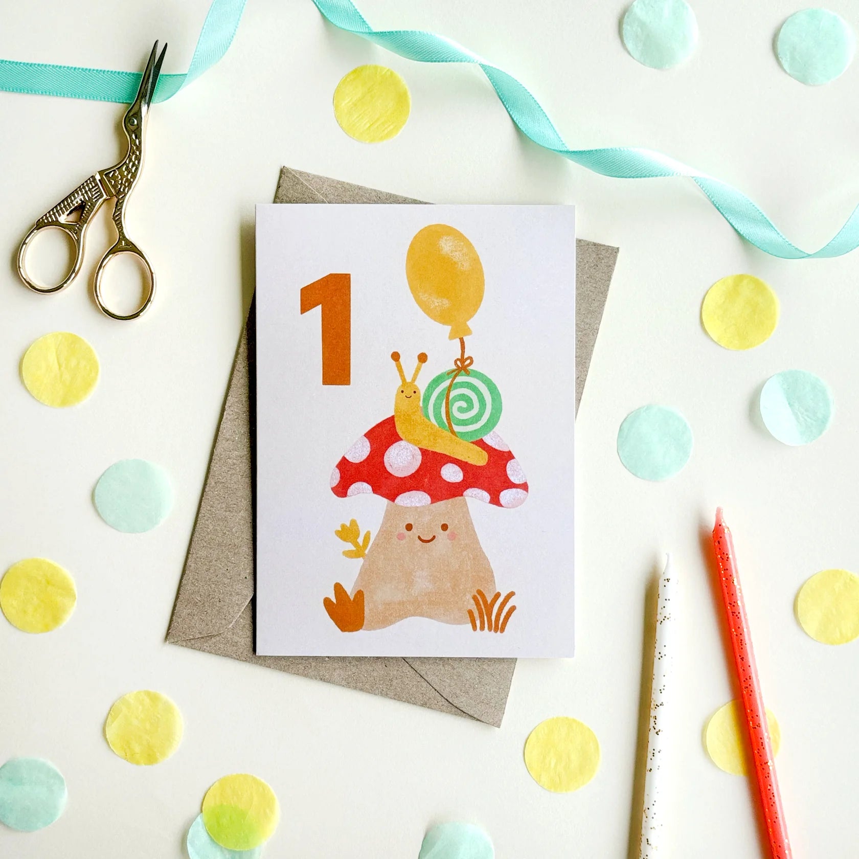 The 1st Birthday Age Milestone Card featuring an illustrated snail with a bright balloon tied to its shell, perched on top of a sweet spotty mushroom A bold hand-lettered number 1 sits on a white background - the colour palette makes it a gender neutral greeting card. The card is surrounded by yellow and blue circle paper cut outs and a pair of scissors and pencils.