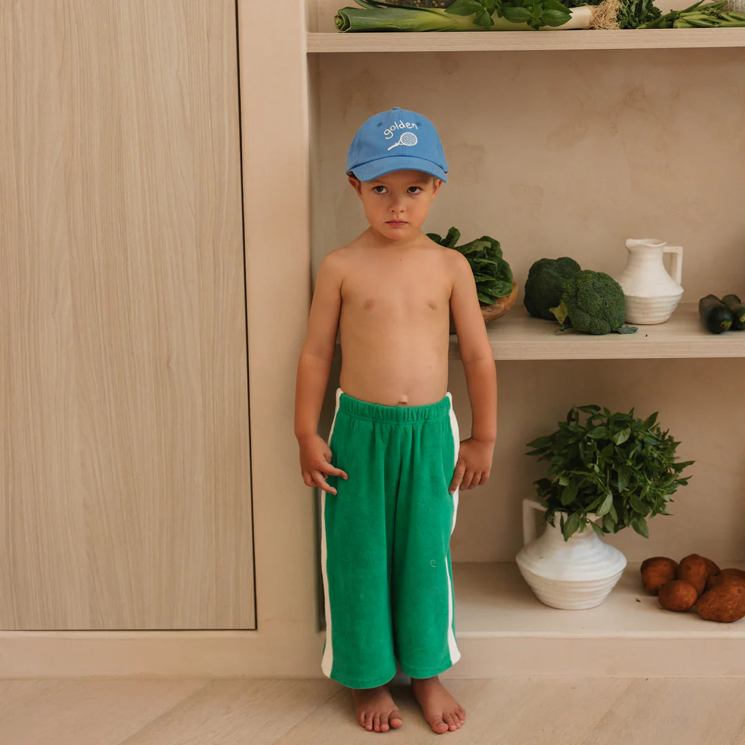 A young boy wears GOLDEN CHILDREN's Ciao Tutti Relaxed Pants Terry Towel Leafy Green, standing barefoot indoors before shelves with vegetables and a white pitcher, looking at the camera with a neutral expression and blue cap.
