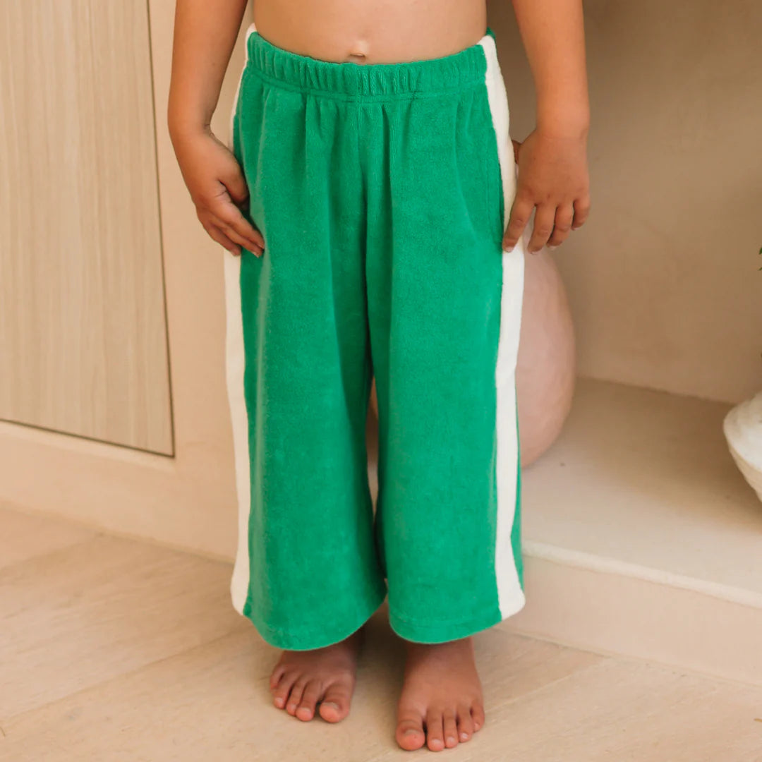 A child stands barefoot on a light floor wearing GOLDEN CHILDREN's "Ciao Tutti Relaxed Pants Terry Towel Leafy Green," featuring bright green wide legs, white side stripes, and an elastic waistband. The upper body and face are not visible.