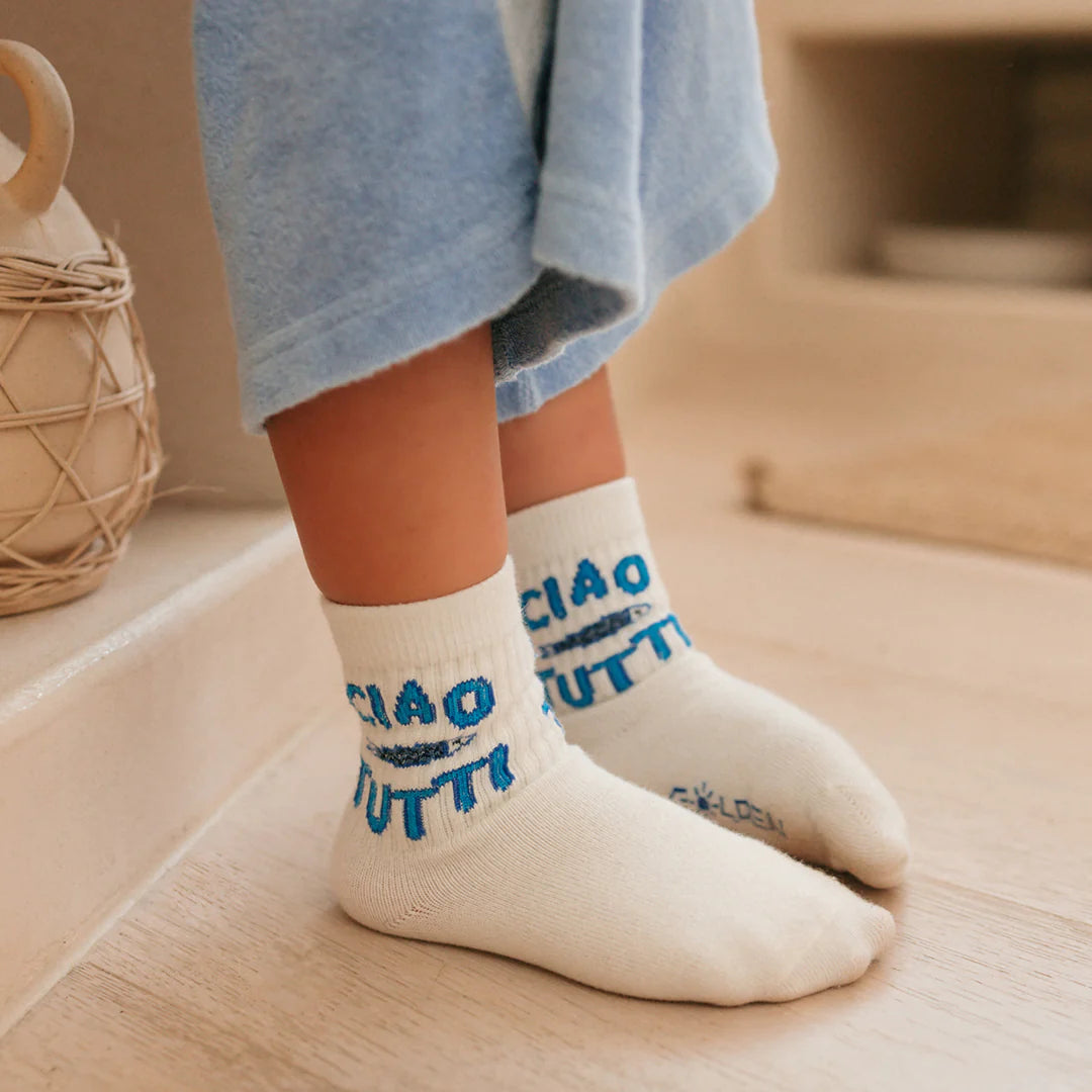 A child in light blue pants wears GOLDEN CHILDREN Ciao Tutti Pesci Socks Vanilla with reinforced toe caps, standing on a light wooden floor near a woven basket.