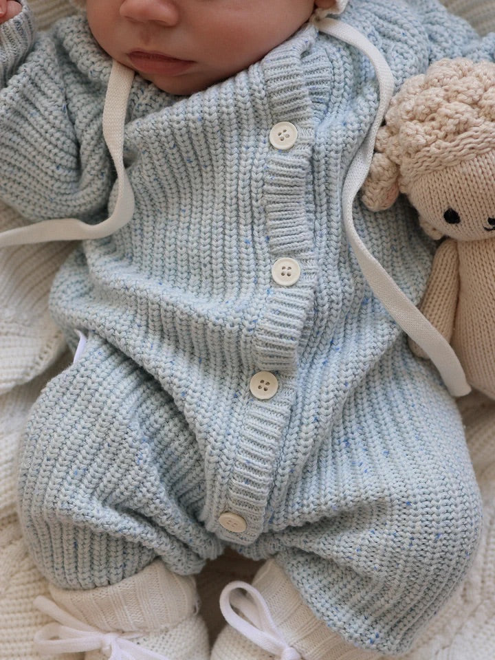 A baby in a ZIGGY LOU Classic Knit Romper Aero Fleck with white buttons lies down, holding a beige knitted stuffed animal. Only the lower half of the face and body are visible.