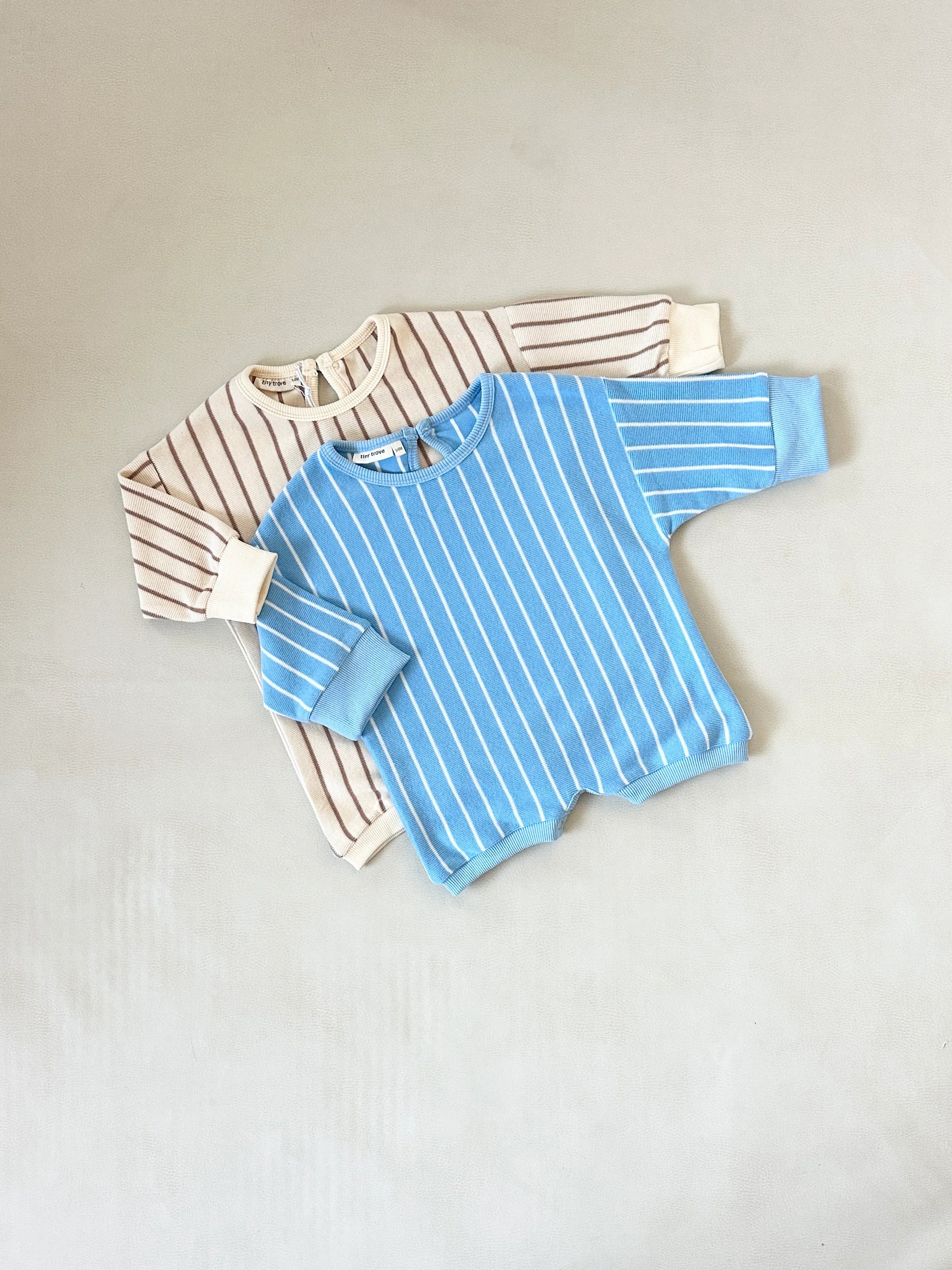 Two Tiny Trove Addie Cotton Stripe Rompers (one beige/brown stripe, one blue/white stripe) from TINY TROVE, made of 100% cotton with ribbed cuffs and relaxed fit, are displayed on a light surface.