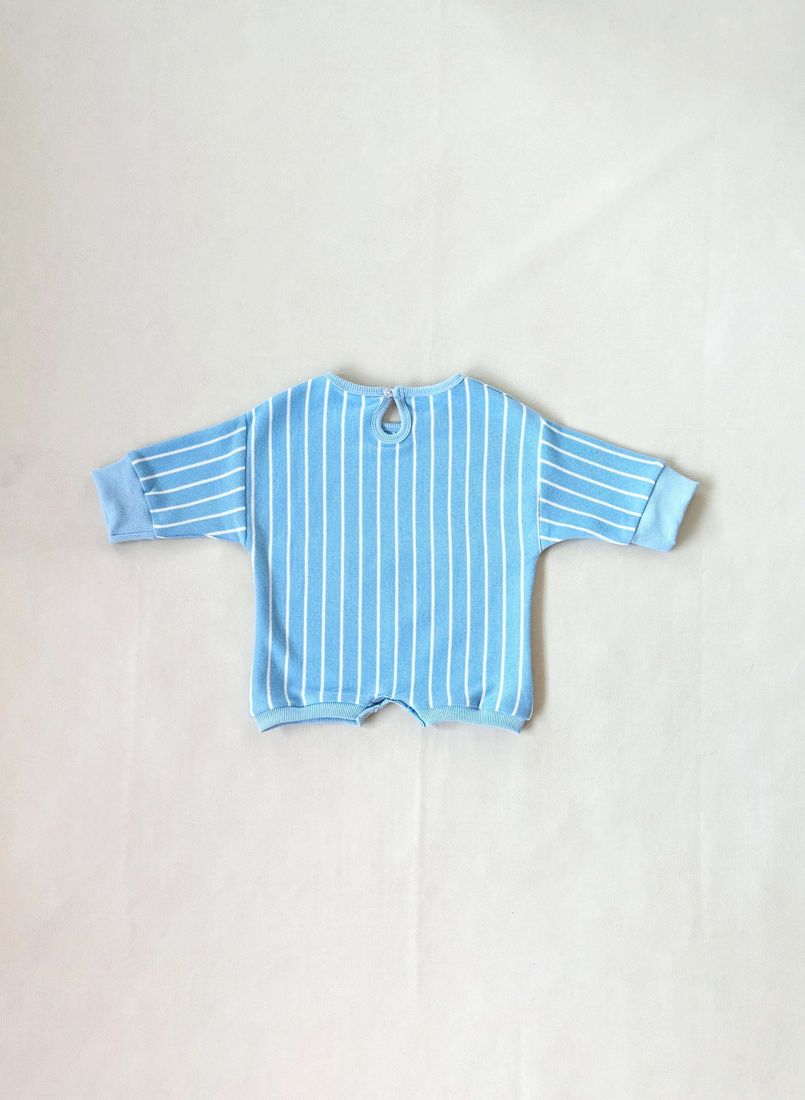 The TINY TROVE Tiny Trove Addie Cotton Stripe Romper in blue/white stripe features long sleeves, 100% cotton fabric, white vertical stripes, wide cuffs, and a buttoned keyhole back, shown laid flat on a neutral background.