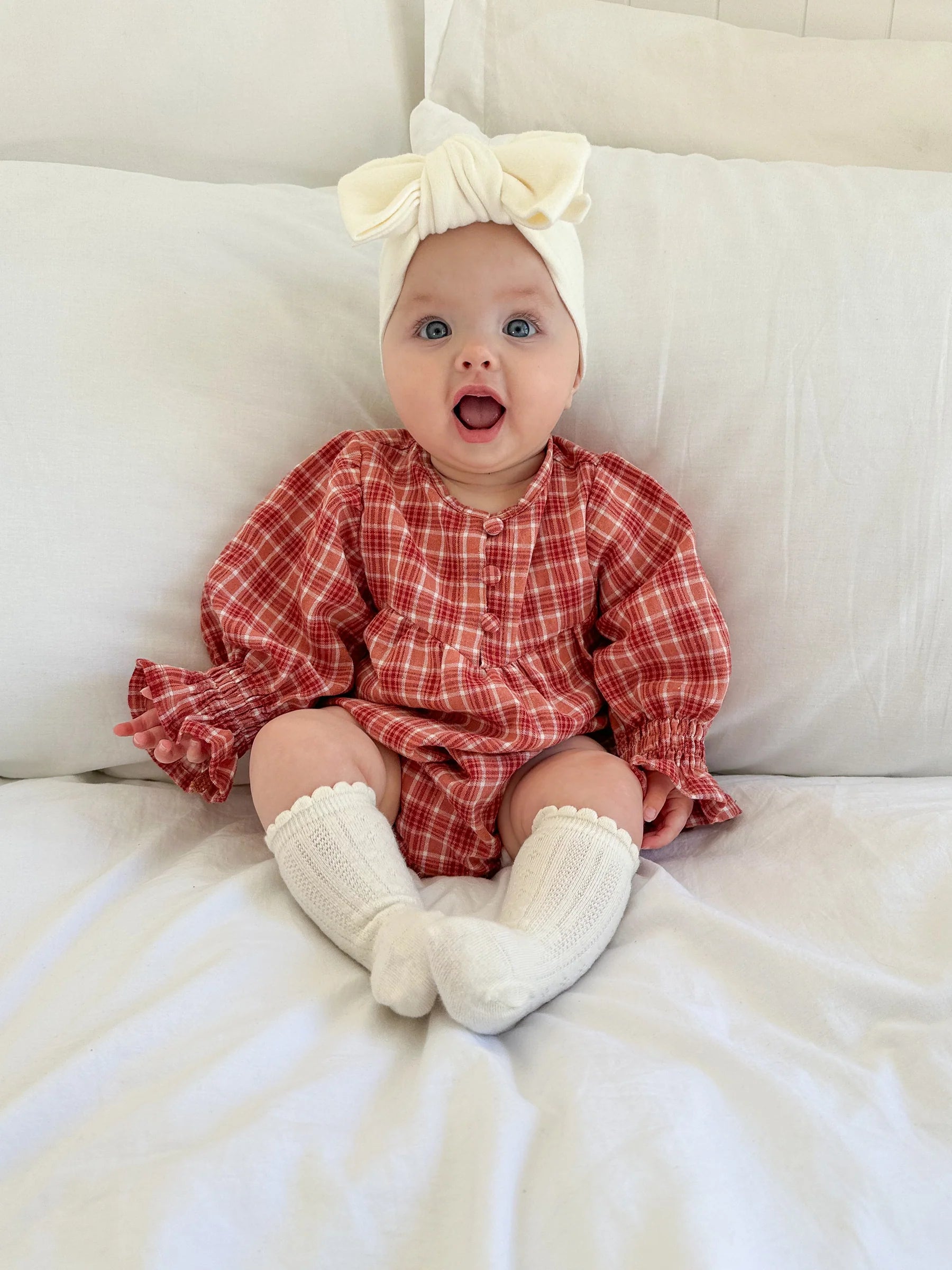 A blue-eyed baby in a red plaid dress and white knee-high socks sits safely on a white bed, wearing a large Ziggy Lou - Headband Milk by ZIGGY LOU, with a surprised expression.