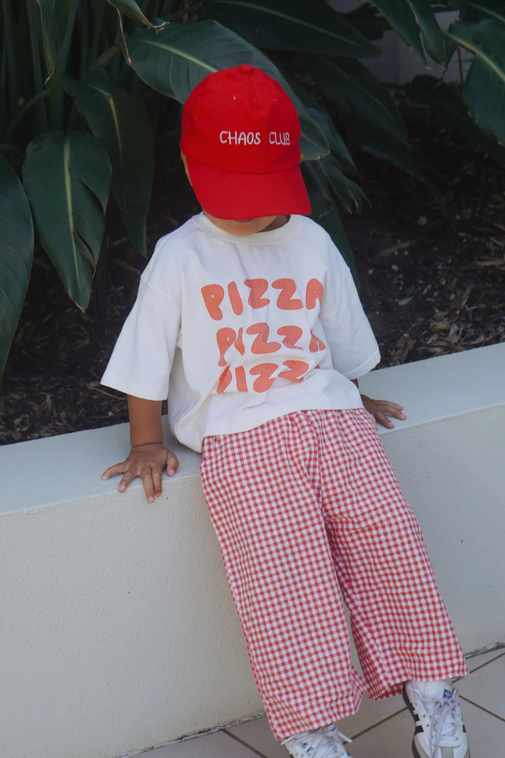 A young child in a red "Chaos Club" cap, white "PIZZA PIZZA PIZZA" tee, WORN CURATIONS' All Day Pant Chilli with an elastic waistband, and white sneakers leans against a low wall, lush green leaves behind.