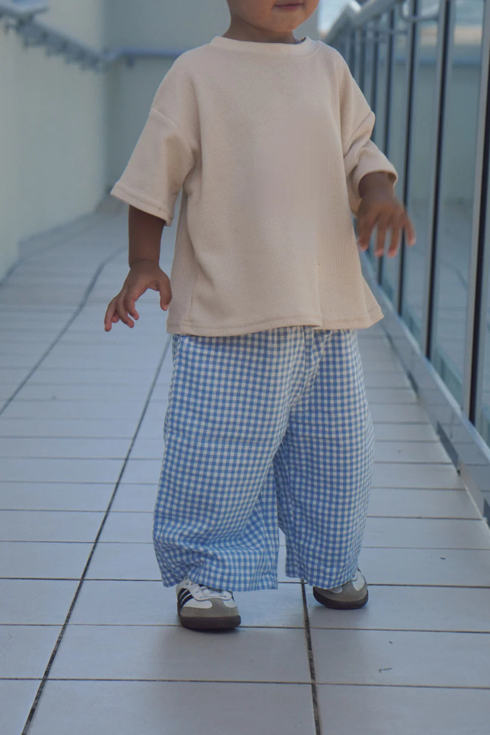 A young child stands on a tiled walkway in a cream oversized shirt, sneakers, and WORN CURATIONS All Day Pant Cornflower. The child's face is partially out of frame.