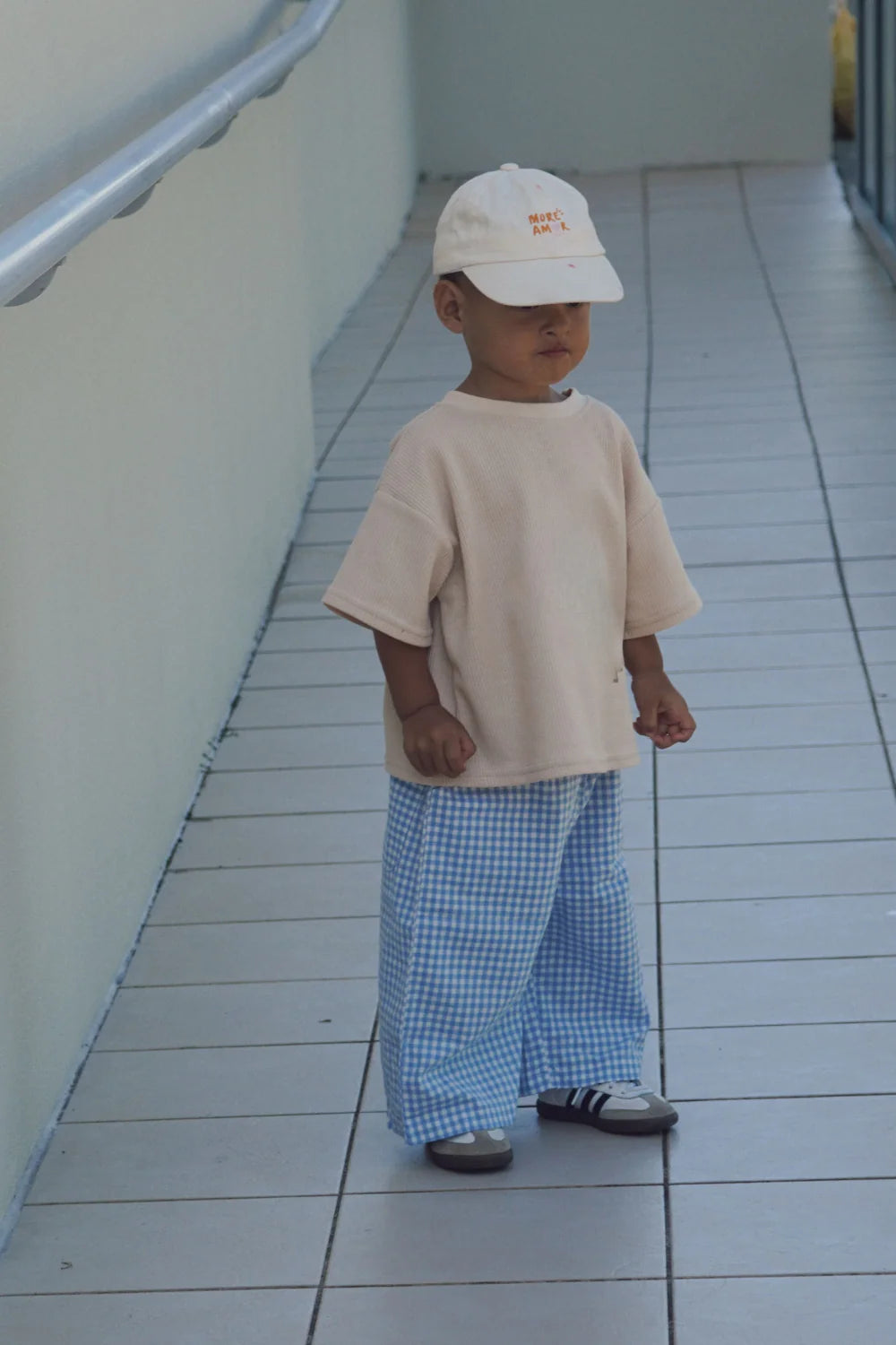 A young child stands on a tiled walkway wearing a cream shirt, white cap, sneakers, and WORN CURATIONS All Day Pant Cornflower. The cotton wide-leg pants add comfort and style as the child faces forward with a neutral expression.