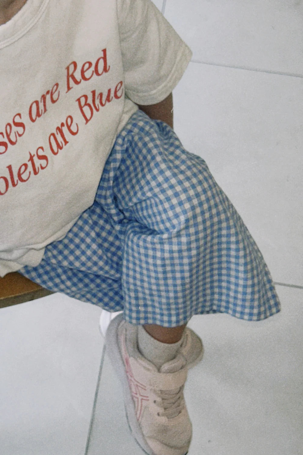 A child sits cross-legged on a tiled floor, wearing a white T-shirt with red text, light pink sneakers, and WORN CURATIONS' Worn Curations - All Day Pant Cornflower—perfect everyday kids’ pants for comfort and style.