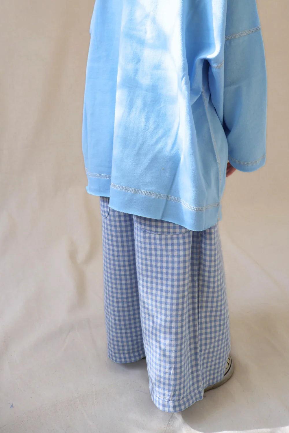 A person stands on a light surface, showing only their legs in WORN CURATIONS' All Day Pant Cornflower, featuring wide-legged blue-and-white gingham checks.