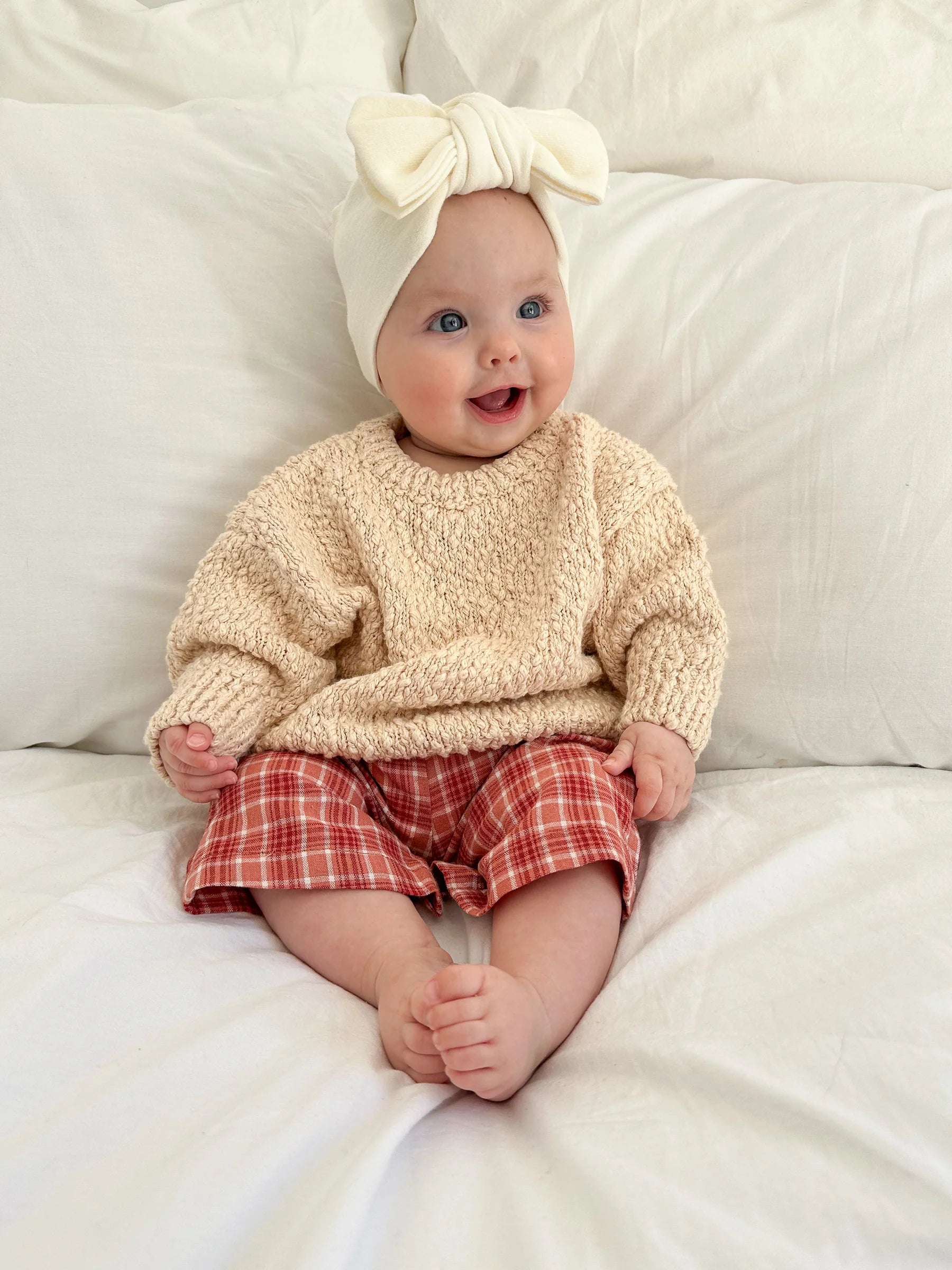 A smiling baby wears a cream knit sweater, red plaid pants, and a Ziggy Lou - Headband Milk by ZIGGY LOU from the Milk Signature Collections while sitting on a white bed with pillows, looking slightly to the side.