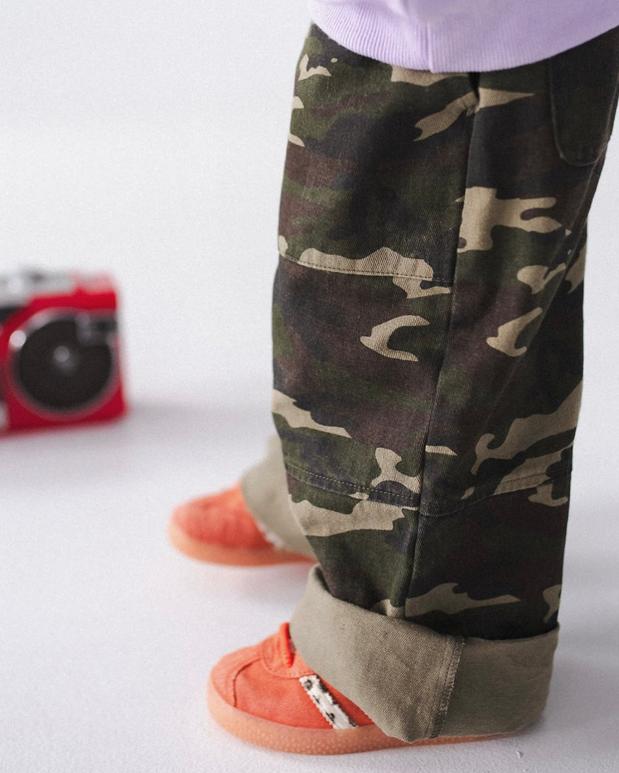 A person wears SAM THE LABEL’s Sam the Label - The Cargo Pant Camo and orange sneakers, standing on a white surface with a red portable speaker in the background.