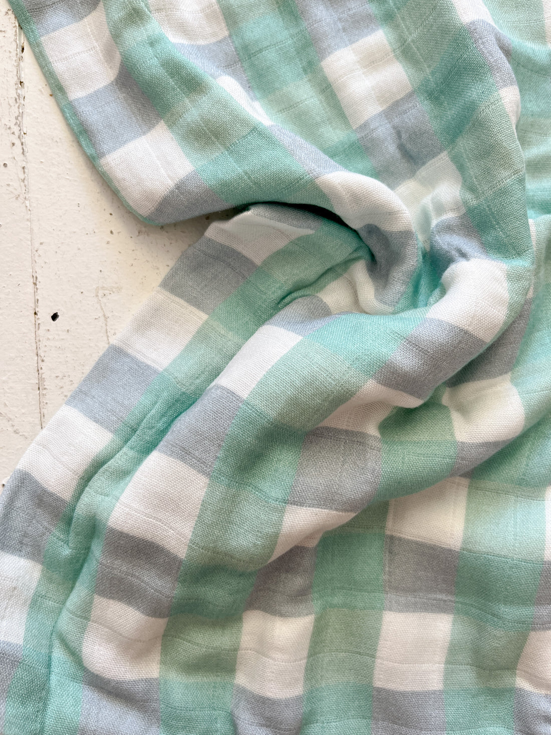 Close-up of the crumpled Milky Designs ~ Seamist Gingham Wrap by MILKY DESIGNS, showcasing a plaid pattern in turquoise, gray, and white. The buttery softness and folds create a casual appearance against a light background.