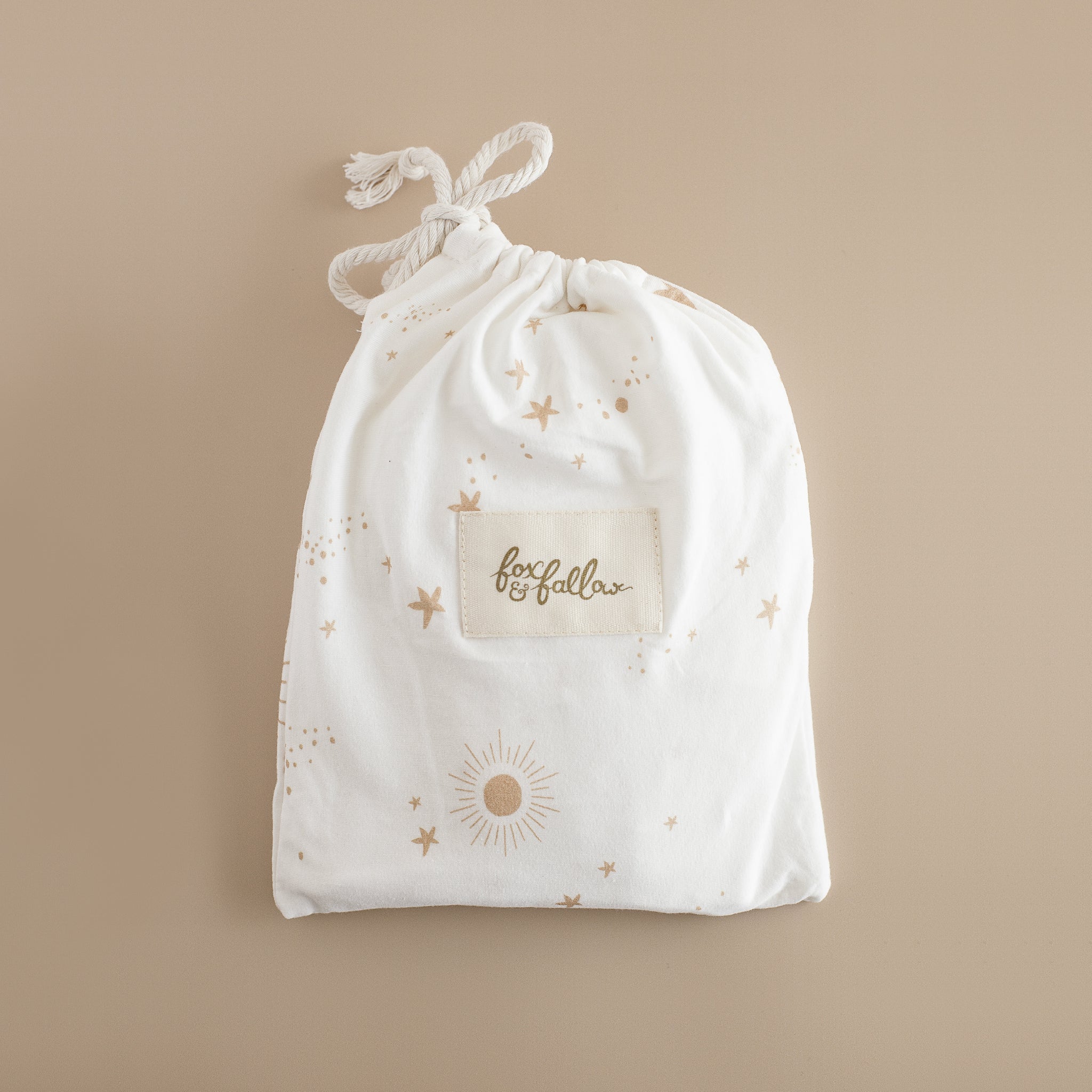 A white drawstring bag with gold star and sun designs, labeled FOX & FALLOW, holds the Constellation Organic Fitted Bassinet Sheet/Change Pad Cover—ideal for storing GOTS certified baby essentials.