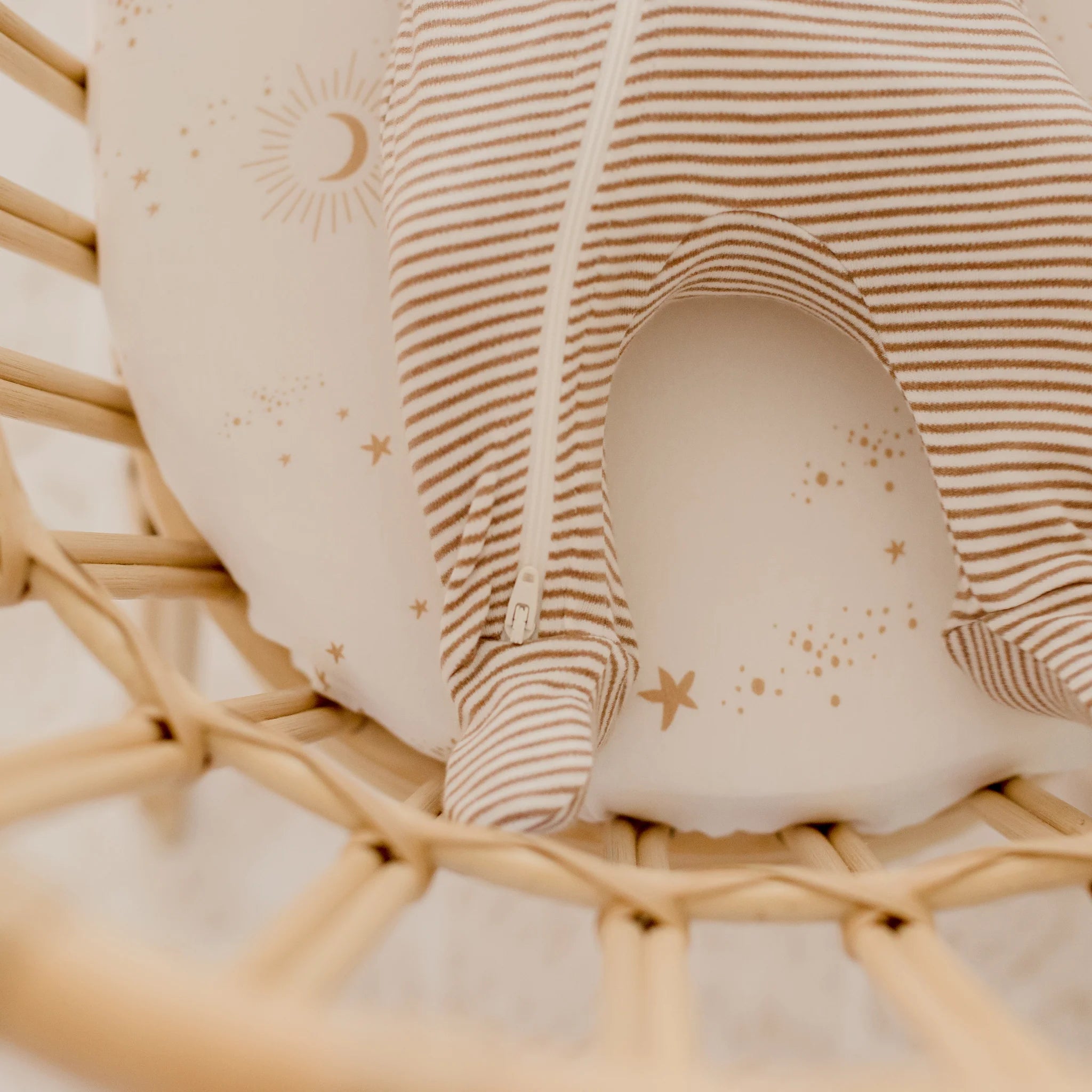 A baby in a brown and white striped onesie lies on the FOX & FALLOW Constellation Organic Fitted Bassinet Sheet/Change Pad Cover, featuring golden stars, moons, and suns, inside a wicker bassinet.