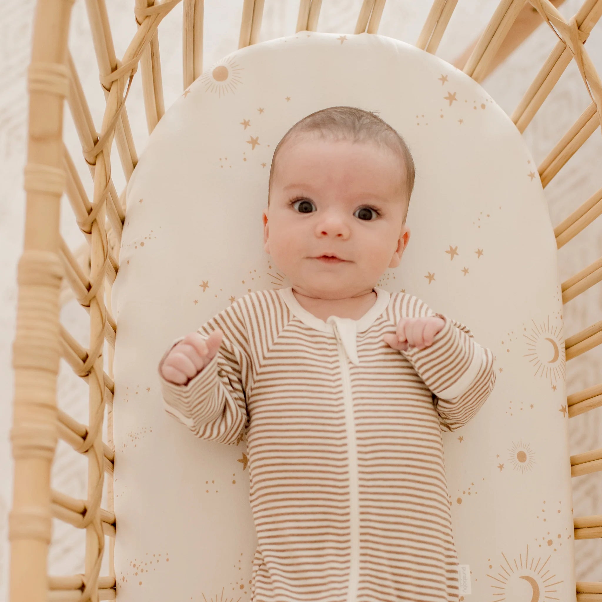 A baby lies on their back in a woven bassinet with FOX & FALLOW's Constellation Organic Fitted Bassinet Sheet, featuring a sun and star pattern in soft neutral tones. The bedding is GOTS certified for quality and comfort.