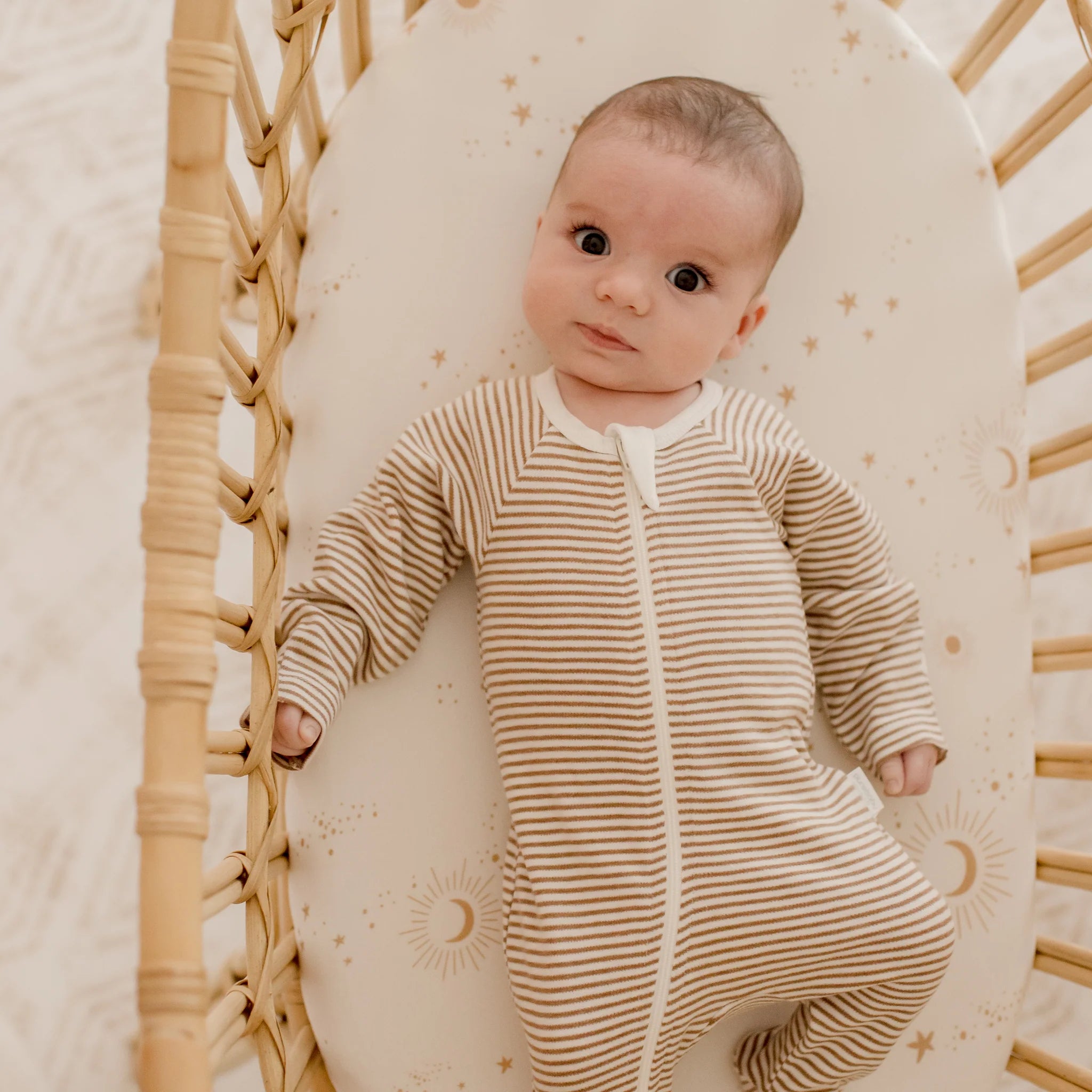 A baby lies on a woven bassinet, gazing up with wide eyes, resting on the FOX & FALLOW Constellation Organic Fitted Bassinet Sheet featuring sun, moon, and star designs for dreamy comfort.