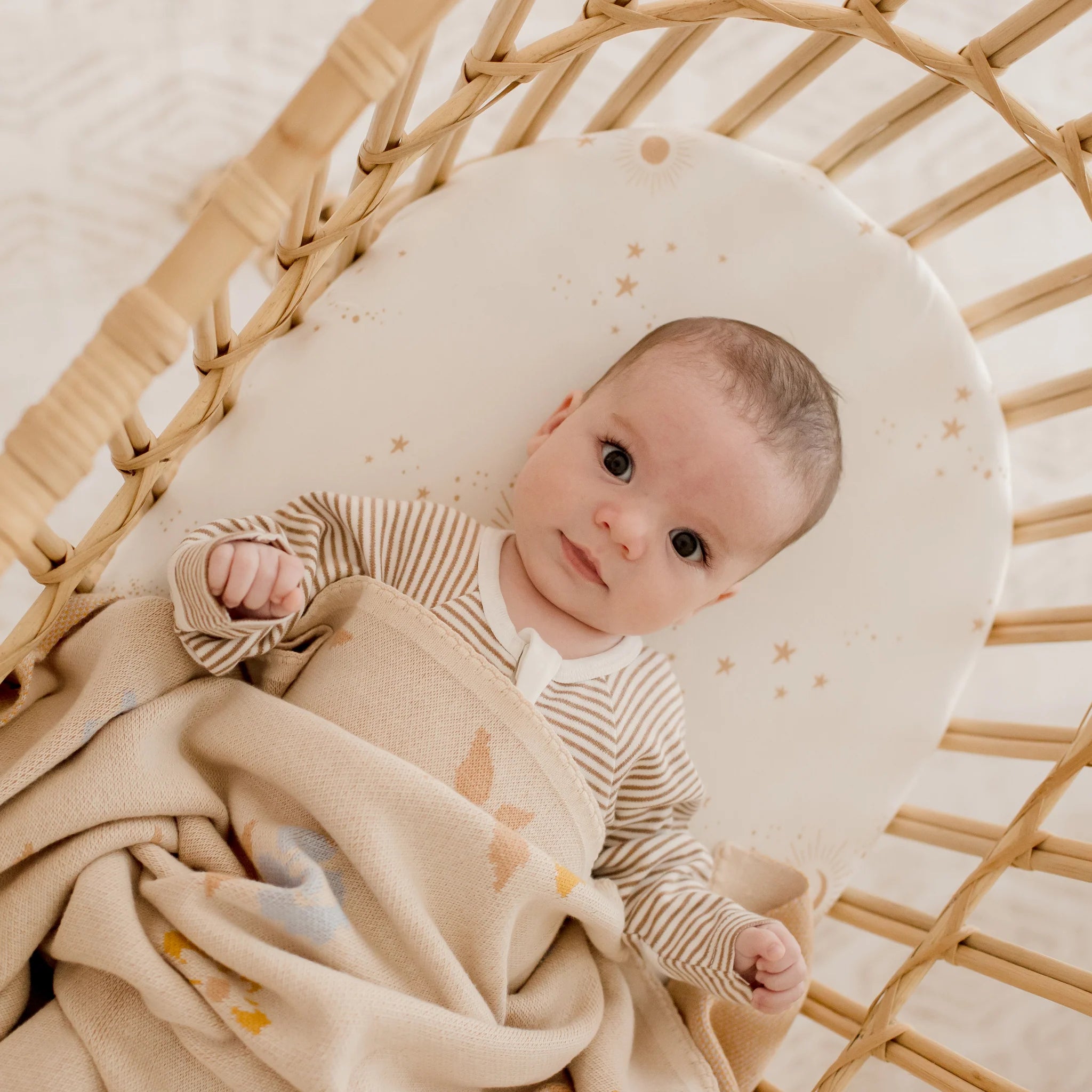 A baby lies in a wicker bassinet, wrapped in a beige blanket and wearing a striped onesie, on the FOX & FALLOW Fox & Fallow - Constellation Organic Fitted Bassinet Sheet/Change Pad Cover with a celestial stars and moons pattern.