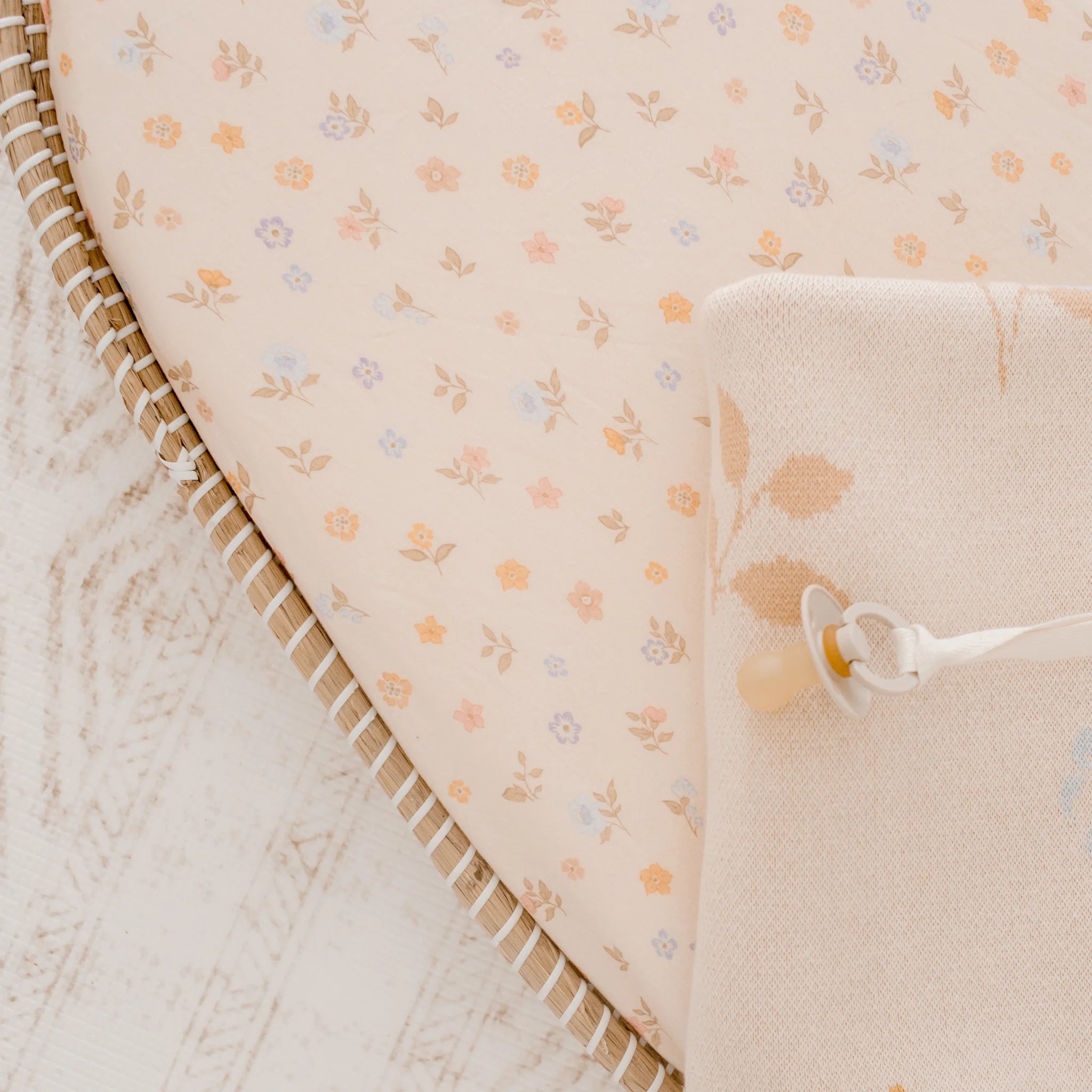A close-up of a baby bassinet with a FOX & FALLOW - Meadow Organic Fitted Bassinet Sheet/Change Pad Cover in a floral print, a beige blanket and white pacifier, all placed on a light wooden floor.