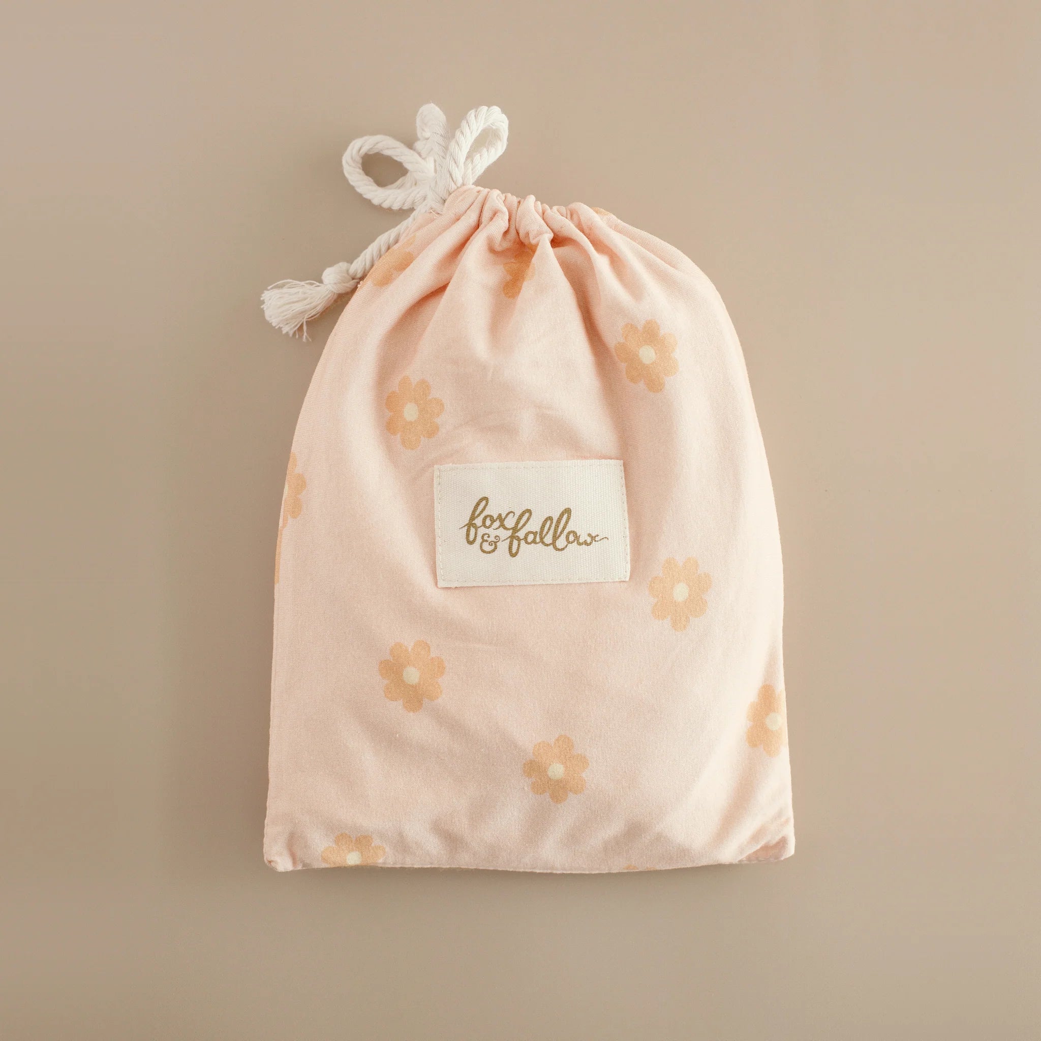 A pale pink FOX & FALLOW drawstring bag with light orange daisies and a white "fox & fallow" label lies on a beige surface, perfect for gifting the Daisy Chain Organic Fitted Bassinet Sheet/Change Pad Cover.