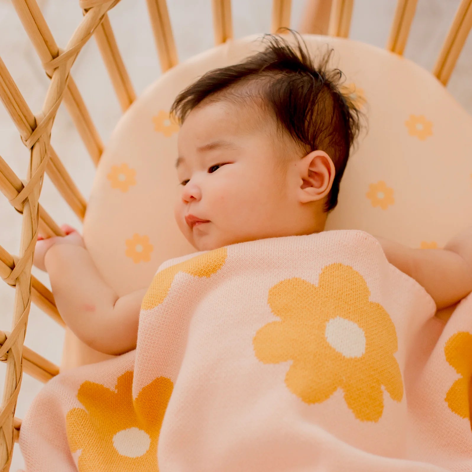 A baby rests calmly in a wooden crib, covered with a soft pink blanket, lying on the FOX & FALLOW Daisy Chain Organic Fitted Bassinet Sheet/Change Pad Cover made from GOTS certified organic materials.