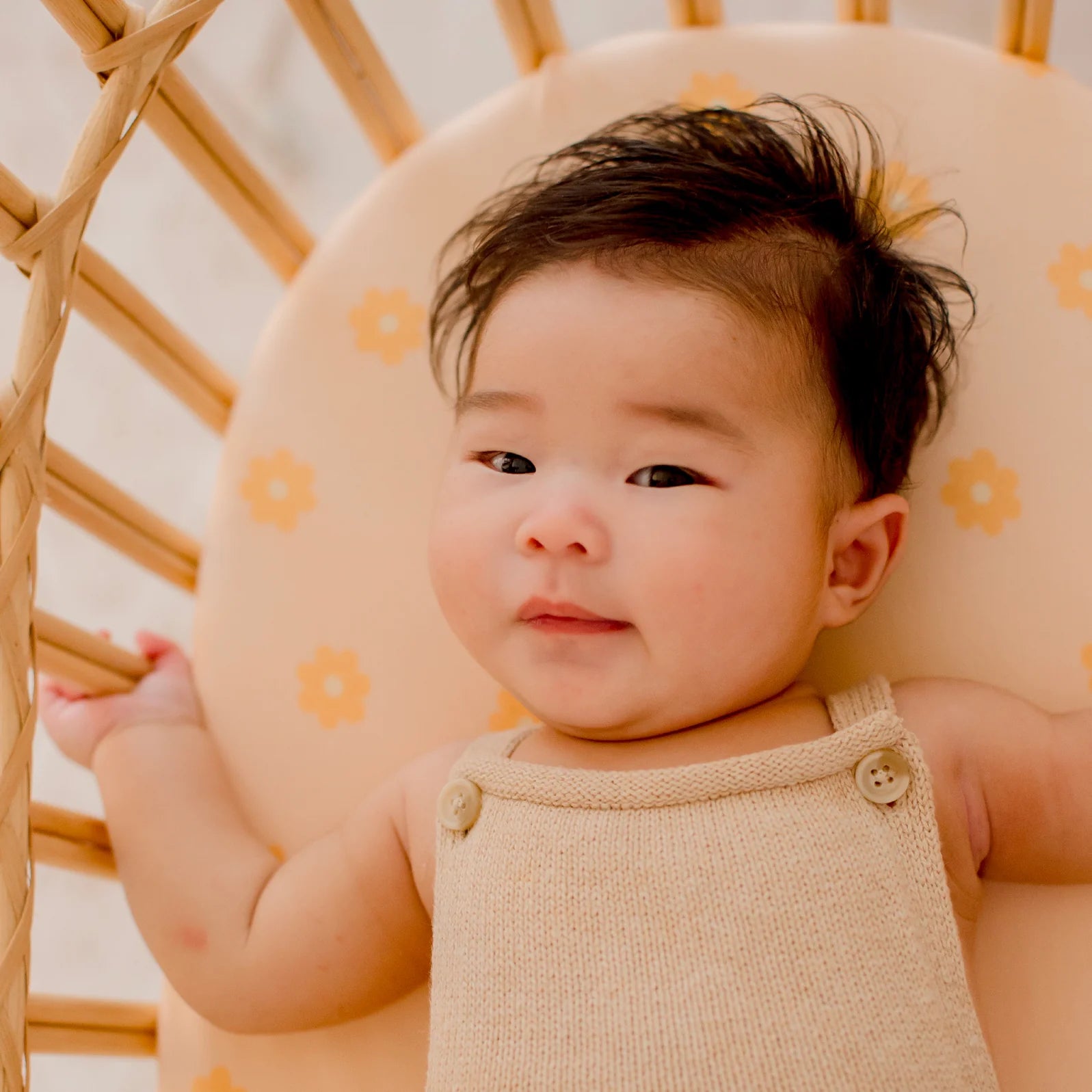A dark-haired baby rests in a wicker bassinet on a FOX & FALLOW Daisy Chain Organic Fitted Bassinet Sheet, featuring light peach tones and yellow floral accents, gazing up calmly.