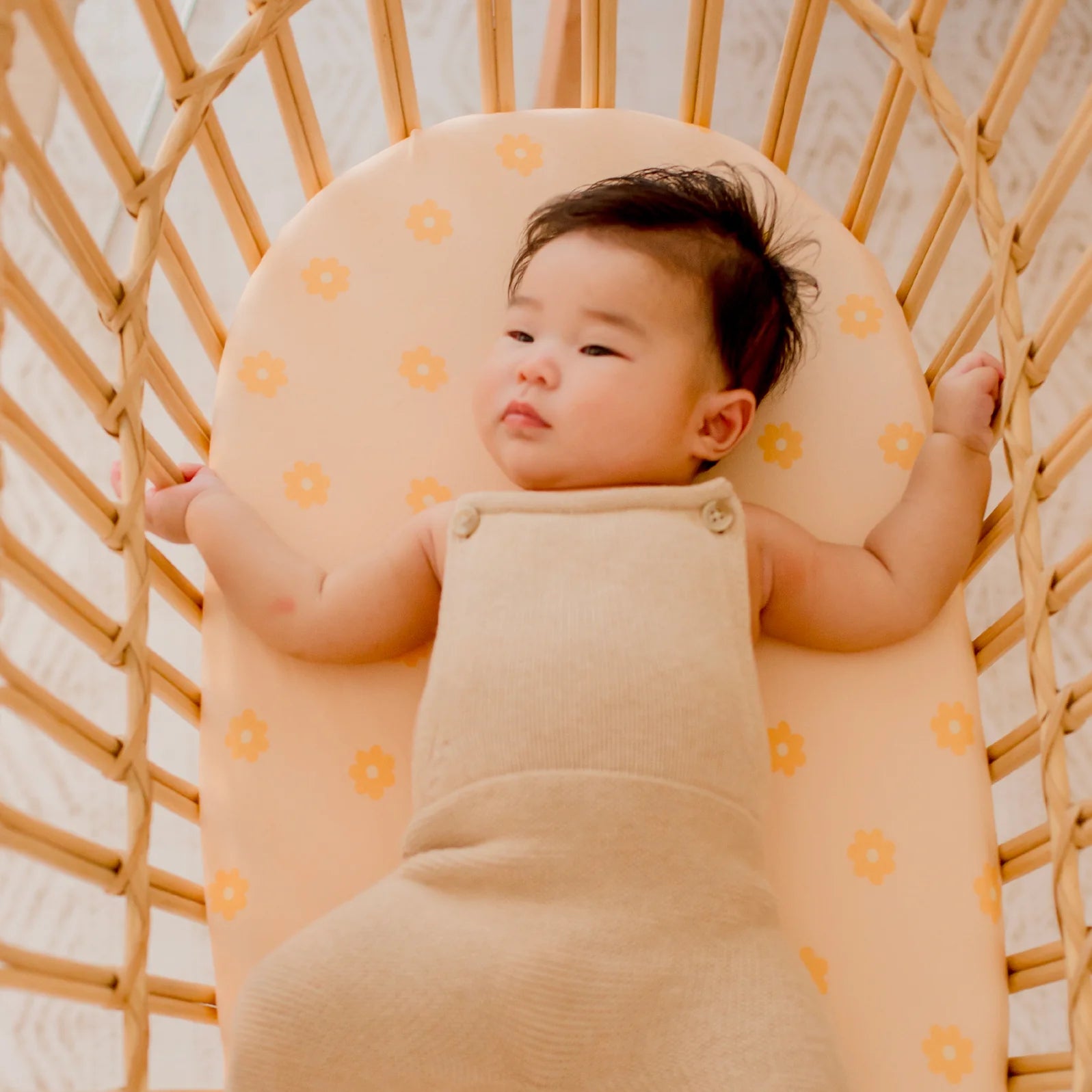 A baby in a beige sleeveless onesie lies on a yellow mattress with floral patterns in a wooden bassinet, resting comfortably on the FOX & FALLOW Daisy Chain Organic Fitted Bassinet Sheet/Change Pad Cover.