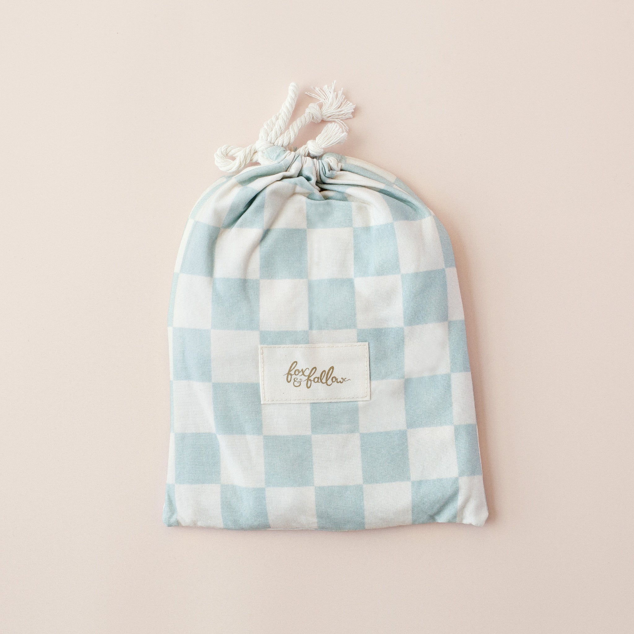 The FOX & FALLOW Sage Check Organic Fitted Bassinet Sheet/Change Pad Cover is shown in a light blue and white checked pouch, making it a stylish gift option for baby bedding essentials.
