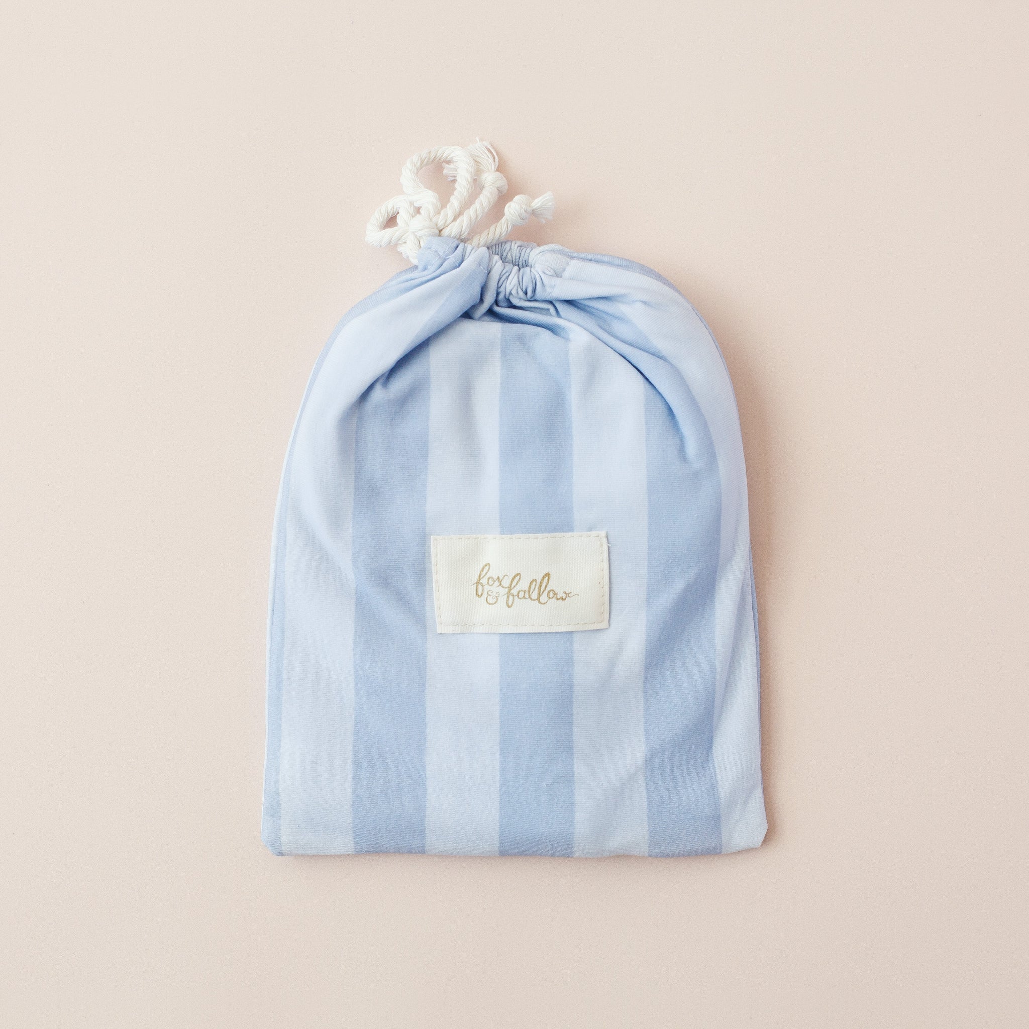 A FOX & FALLOW Cloud Stripe Organic Fitted Bassinet Sheet/Change Pad Cover in light blue and white stripes with a chic “fox follies” label is displayed flat on a pale pink background—a stylish, GOTS certified baby bedding choice.