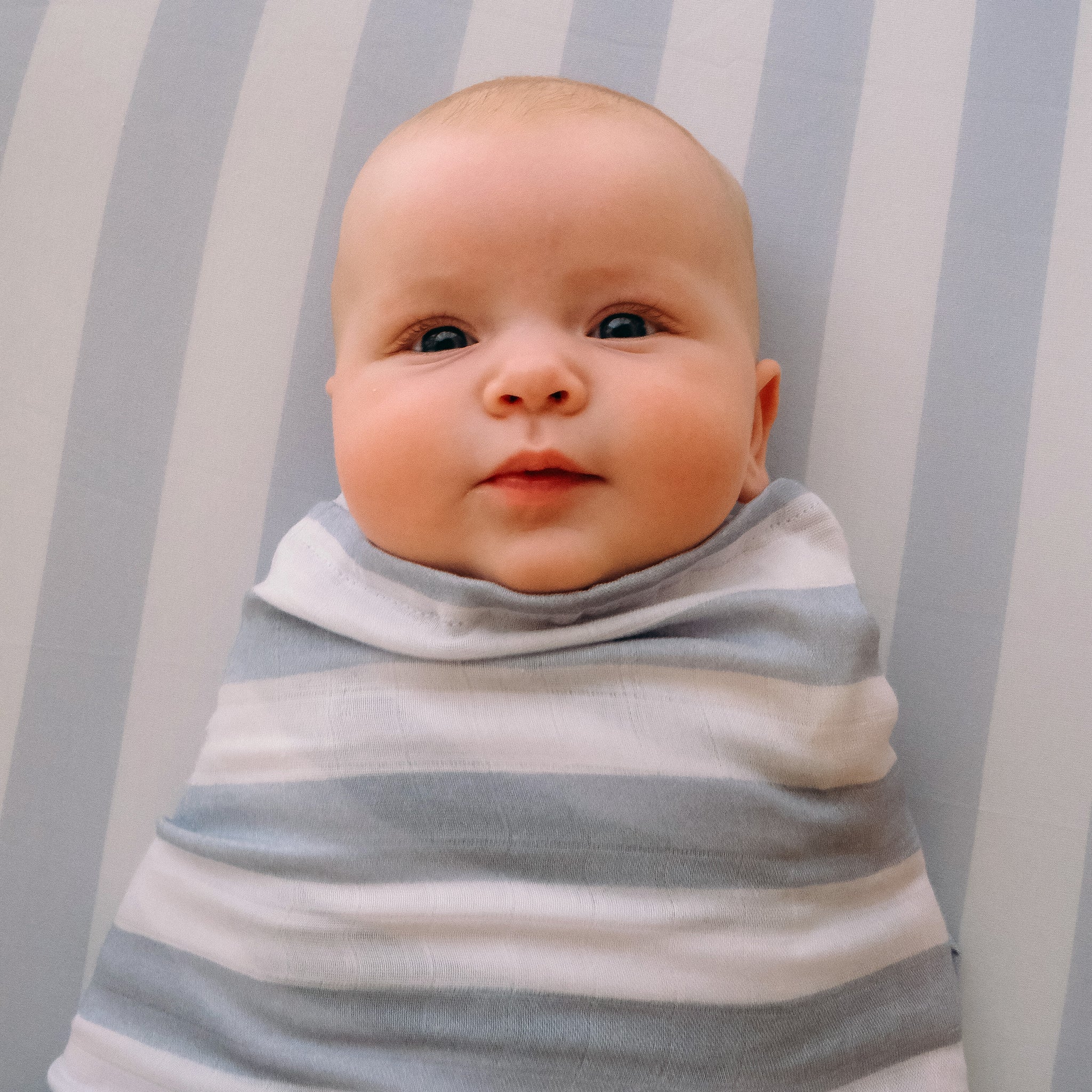 A baby lies on the FOX & FALLOW Cloud Stripe Organic Fitted Cot Sheet, wrapped snugly and gazing upward with a calm expression.